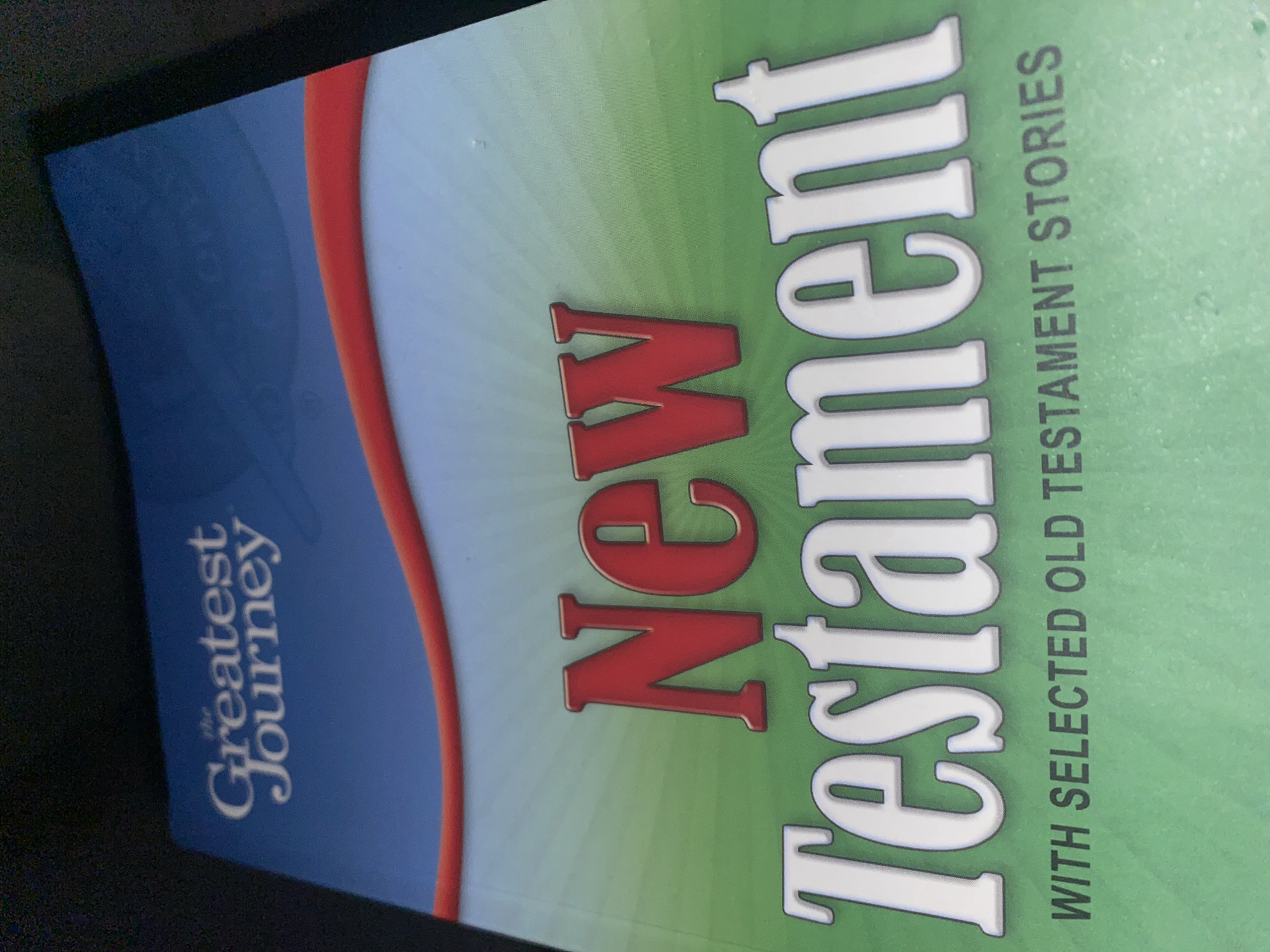 The image shows the cover of a book tilted on its side. The book has a green background with radiating lines. Large white lettering on the left side reads "Testament" and red lettering below it reads "New". To the left of "Testament", in smaller green lettering oriented vertically, are the words "WITH SELECTED OLD TESTAMENT STORIES". On the right side of the book, on a blue background with a red curved stripe, white text reads "the Greatest Journey". The image is a close-up of the book cover, with no discernible city or location details visible.
