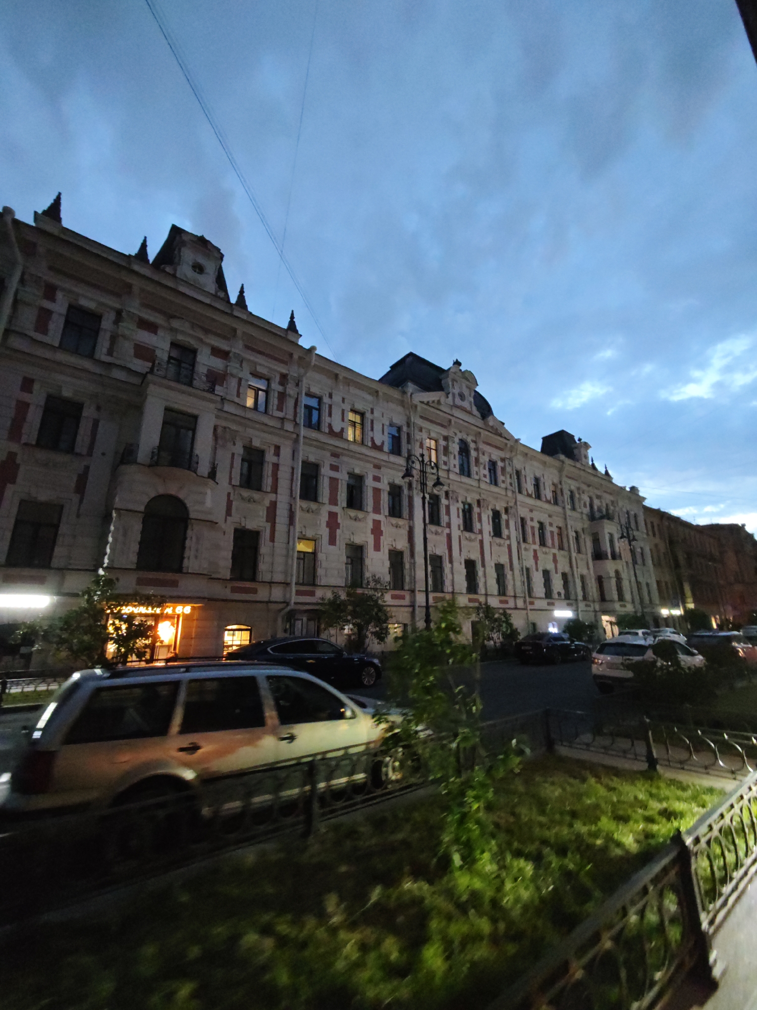 An outdoor, low-angle, wide shot shows a long, multi-story, ornate building in Saint Petersburg, Russia at dusk. The building features a light-colored facade with red decorative elements and many windows. Some windows are illuminated from within. Dark sloped roofs top the building, with decorative spires at the corners. A light pole stands in front of the building. Parked cars, including a silver station wagon, are visible on the street in front of the building. Some of the cars are dark-colored, and at least one is white. A black metal fence runs along the foreground, separating a grassy area from the sidewalk and street. The sky is a deep blue with scattered clouds.