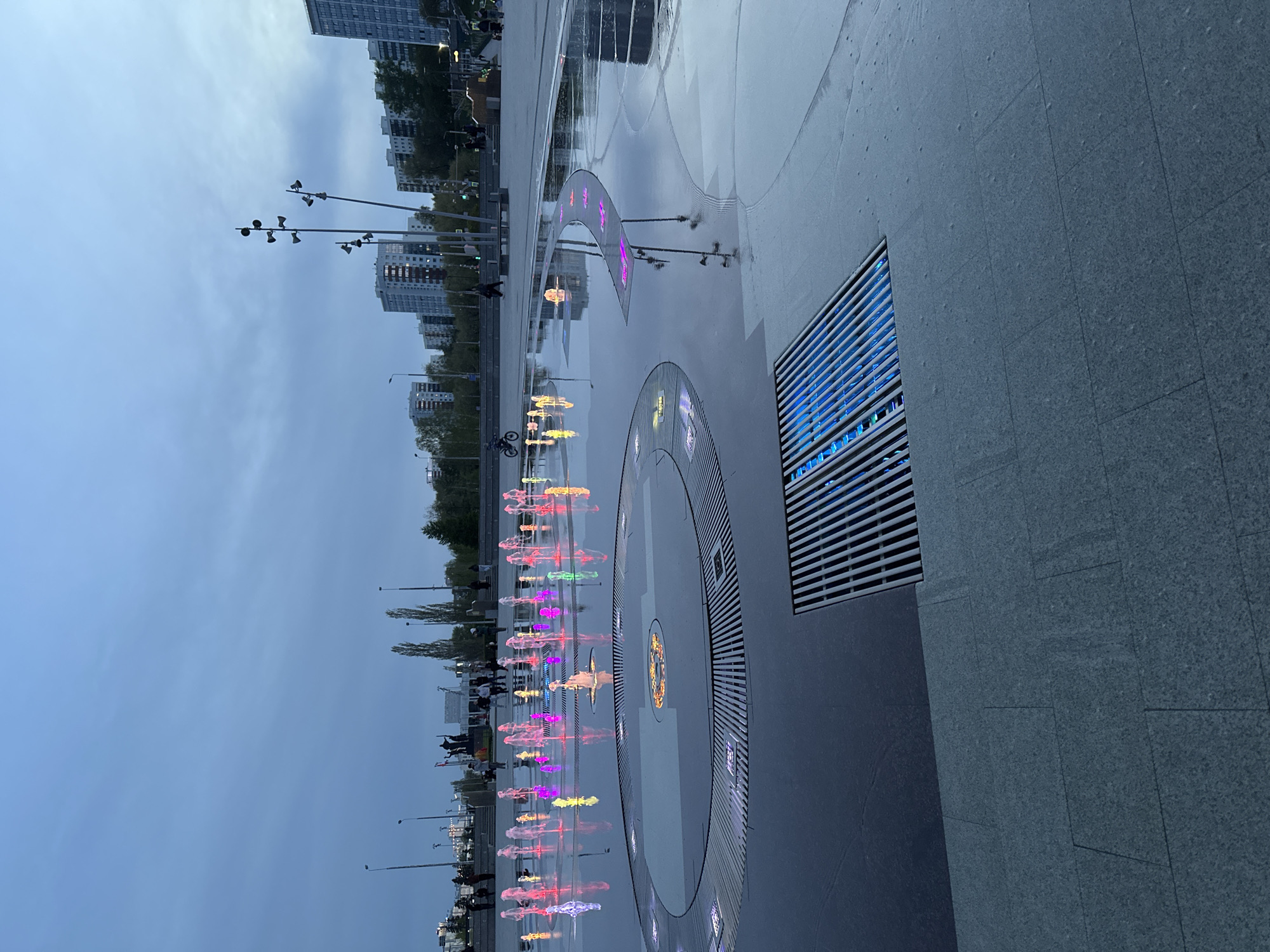 The image captures a scene in Perm, Russia, featuring a modern fountain complex. The foreground is dominated by a large, flat plaza partially covered in shallow water, reflecting the sky and surrounding structures. Integrated into the plaza's surface are circular and curved sections with colorful lights and water jets that create illuminated fountains in shades of red, pink, yellow, and blue. These fountains appear to be a choreographed water show. To the right of the frame is a section of a gray stone wall with a horizontal grate.
In the background, several multistory residential buildings can be seen, and there are tall, thin lampposts with multiple lights that run along the edge of the plaza. People can be seen walking along the perimeter of the fountain, likely enjoying the spectacle. The sky is overcast, suggesting either a late evening or a cloudy day, with a bluish tint due to the lighting conditions. The overall impression is of a modern, urban public space intended for leisure and entertainment.