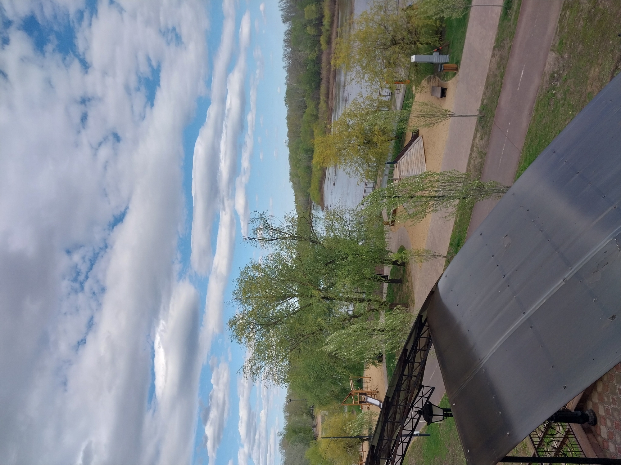 The image shows a view from a building in Orenburg, Russia. The sky is blue with white, puffy clouds. A river is visible in the middle distance, lined with trees. A walkway runs parallel to the riverbank. There is a playground visible among the trees. A portion of a dark, roofed structure is prominent in the foreground.