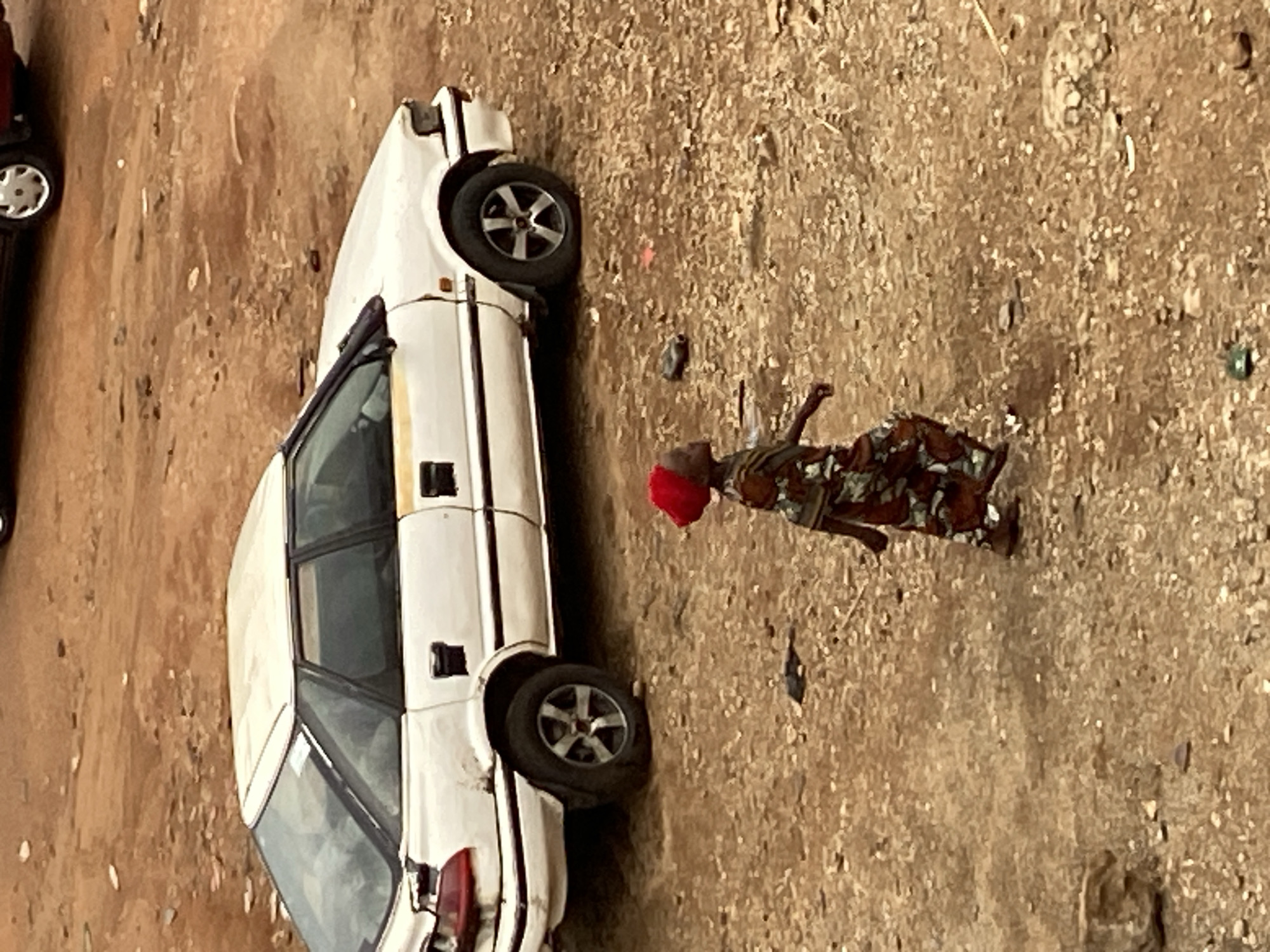 The image shows an outdoor scene, likely a dirt road or unpaved area, in what appears to be Mararaba, Nigeria. The primary subjects are a person and two cars.
A person, possibly a woman, with dark skin and wearing a red head covering, stands upright on the light brown, gravelly ground. She is dressed in a patterned garment, possibly a loose-fitting dress or tunic over pants, with patterns in shades of brown, green, and black. Her right arm is slightly raised, and she appears to be looking towards the foreground vehicle.
To the left of the person is a prominent white four-door vehicle, which resembles an older model sedan or station wagon. It has black-accented wheels, and appears stationary, possibly parked. Its body shows some signs of wear and dirt. In the upper left background, part of another car, dark red or maroon in color, is visible, also with silver wheels.
The ground is dry, unpaved, and covered with small stones and debris, consistent with a rural or semi-urban outdoor setting. The lighting suggests it is daytime, possibly bright and clear, as there are no harsh shadows to indicate early morning or late afternoon. No specific activity beyond standing or observing is clearly depicted for the person. No visible text can be clearly discerned on any object in the image.