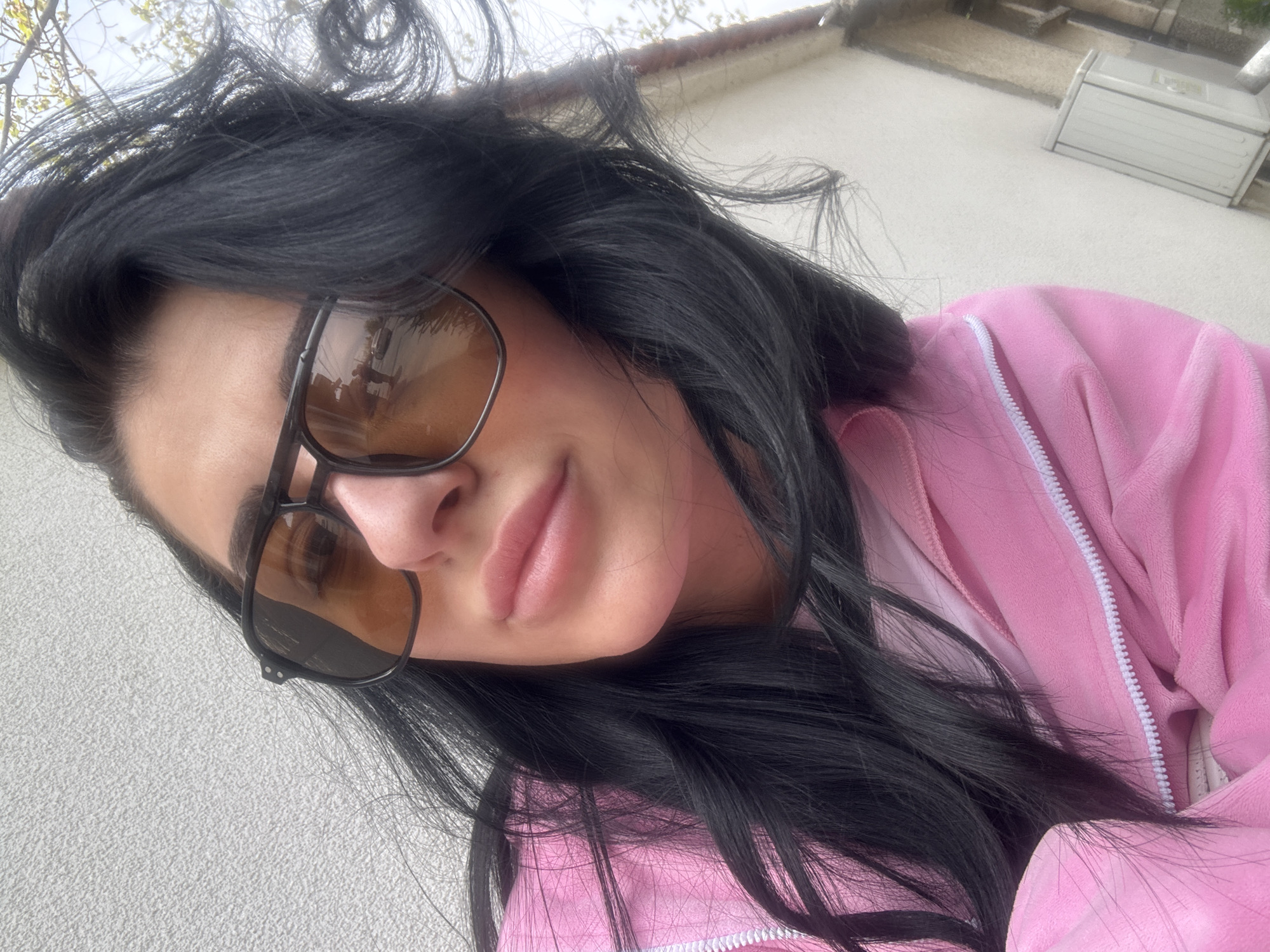 A woman with dark hair and sunglasses is wearing a pink velvet jacket with a white zipper. The background consists of a white wall, part of a building, and some sky with tree branches. The photograph was taken in Saedinenie, Bulgaria.