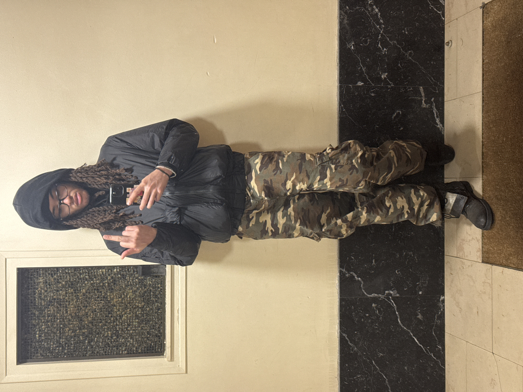 A person wearing a black hooded jacket and camouflage pants stands in a hallway with black and white marble-patterned flooring and beige walls. The person is holding a smartphone and extending their middle finger. There is a window with a textured screen to their left and a brown doormat to their right. The image provides no evidence to confirm the location is Paris, France.