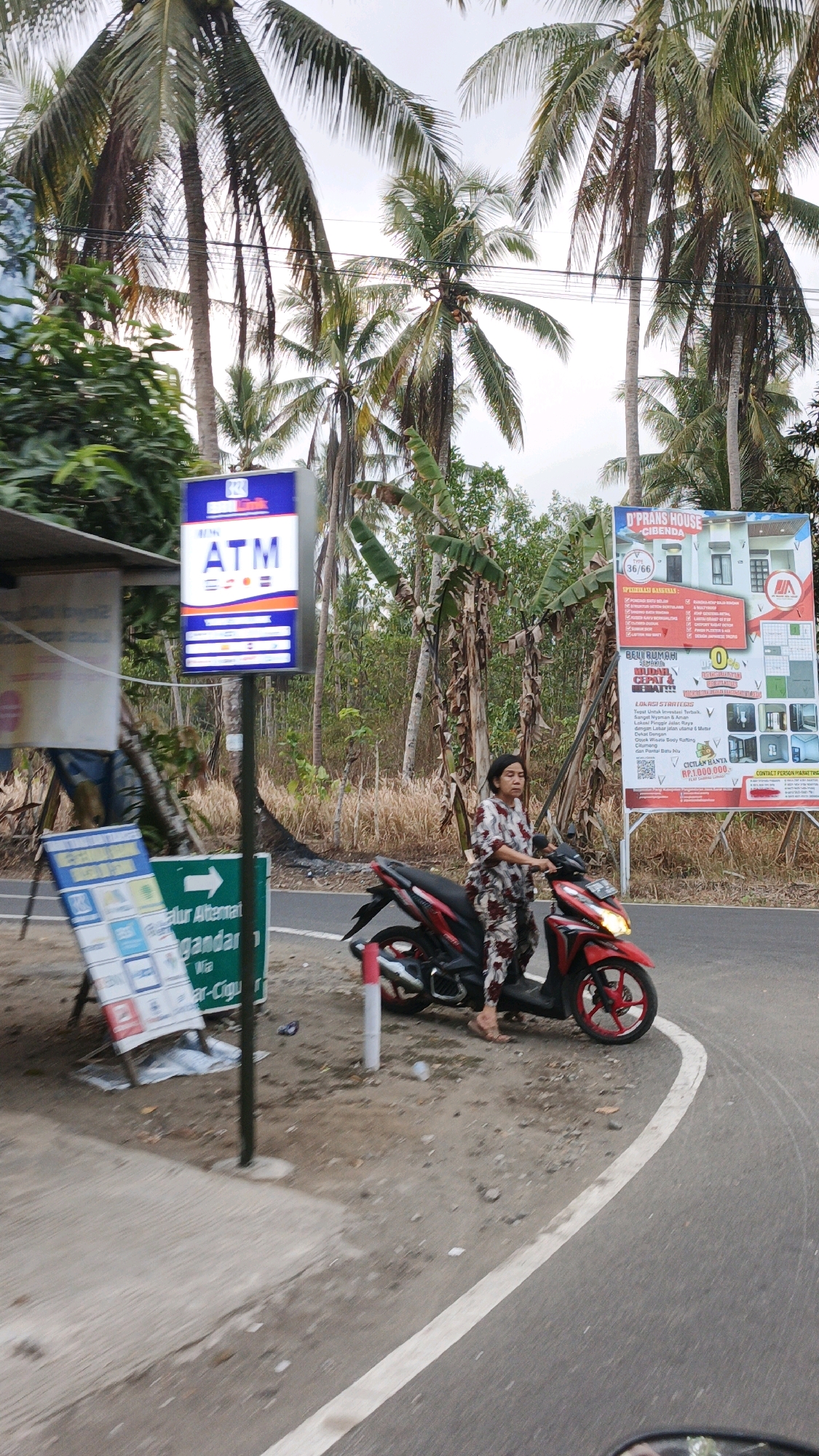 An overcast day in Sukaresik, Indonesia, features a woman in a patterned outfit standing with her bare feet on the ground next to a red and black motorcycle. She is facing the right side of the frame, looking down. The motorcycle is parked on the shoulder of a paved road that curves to the right. To the left of the road, a tall, blue and white ATM sign stands on a pole. A white sign board with multiple logos is leaning on the ground to the left of the ATM sign. In the background, a large billboard advertising "D'PRANS HOUSE CIBENDA" is visible, along with lush green vegetation and numerous palm trees that dominate the skyline.