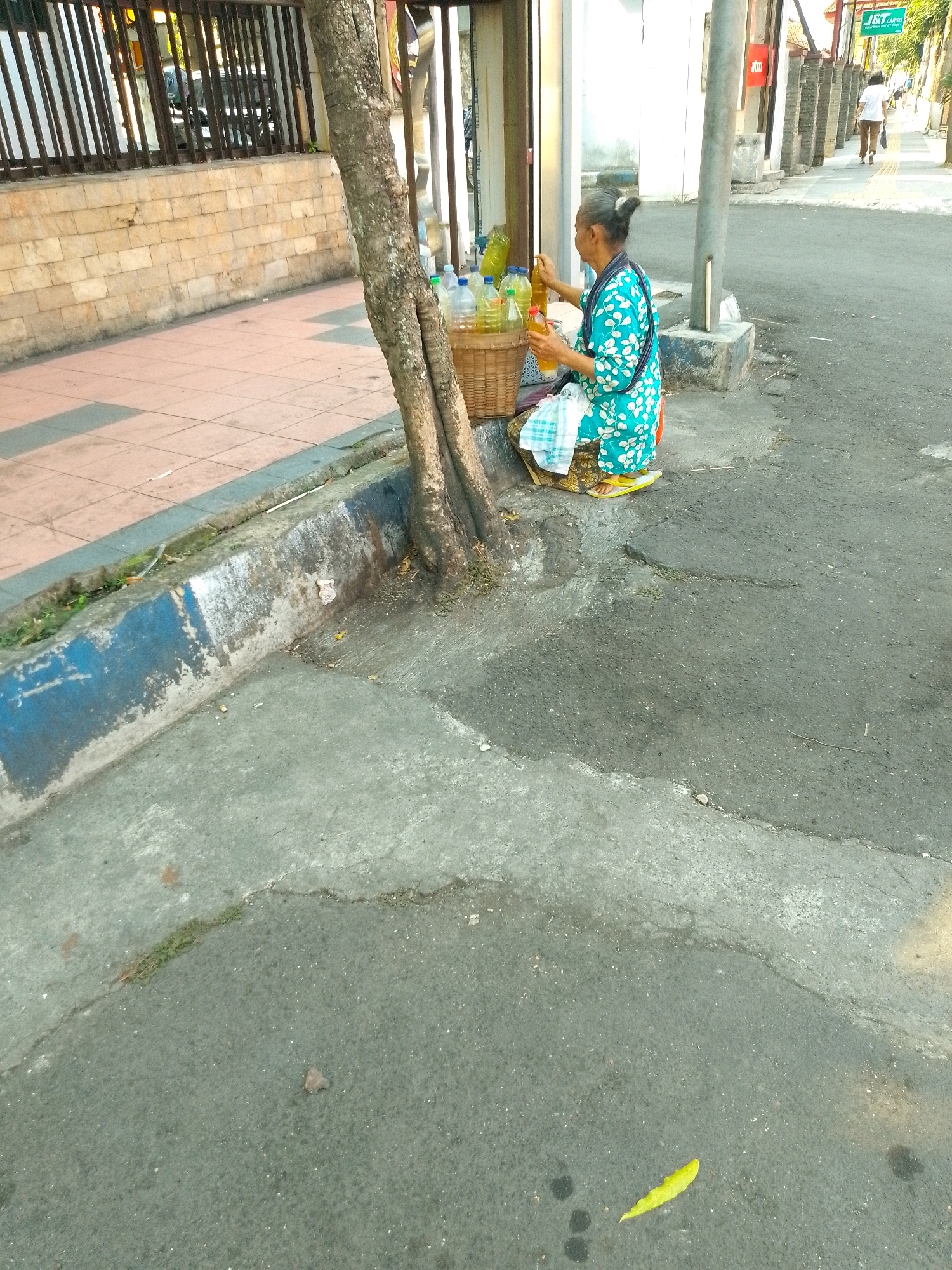 In Jember, Indonesia, an elderly woman sits on the sidewalk next to a tree. She is wearing a blue and white patterned dress and is holding a bottle of clear liquid. In front of her is a woven basket filled with more bottles of the same liquid. The sidewalk is made of light-colored tiles and is bordered by a concrete curb. Behind the woman, there is a building with a metal fence. The street is paved with asphalt and is mostly empty, except for a single yellow leaf on the ground. The lighting suggests it is late afternoon.