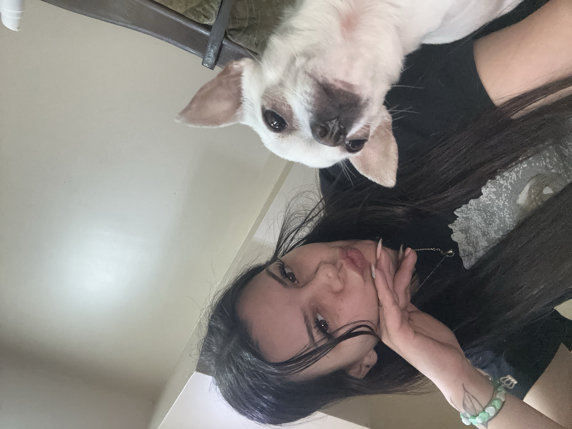 A young woman with long, dark hair poses with a small white dog. The woman's face is turned toward the camera, and her hand is touching her face. She is wearing a dark top. The dog is positioned above her head, looking down at the camera. The background is a light-colored wall and a chair. The location is not discernible from the image.