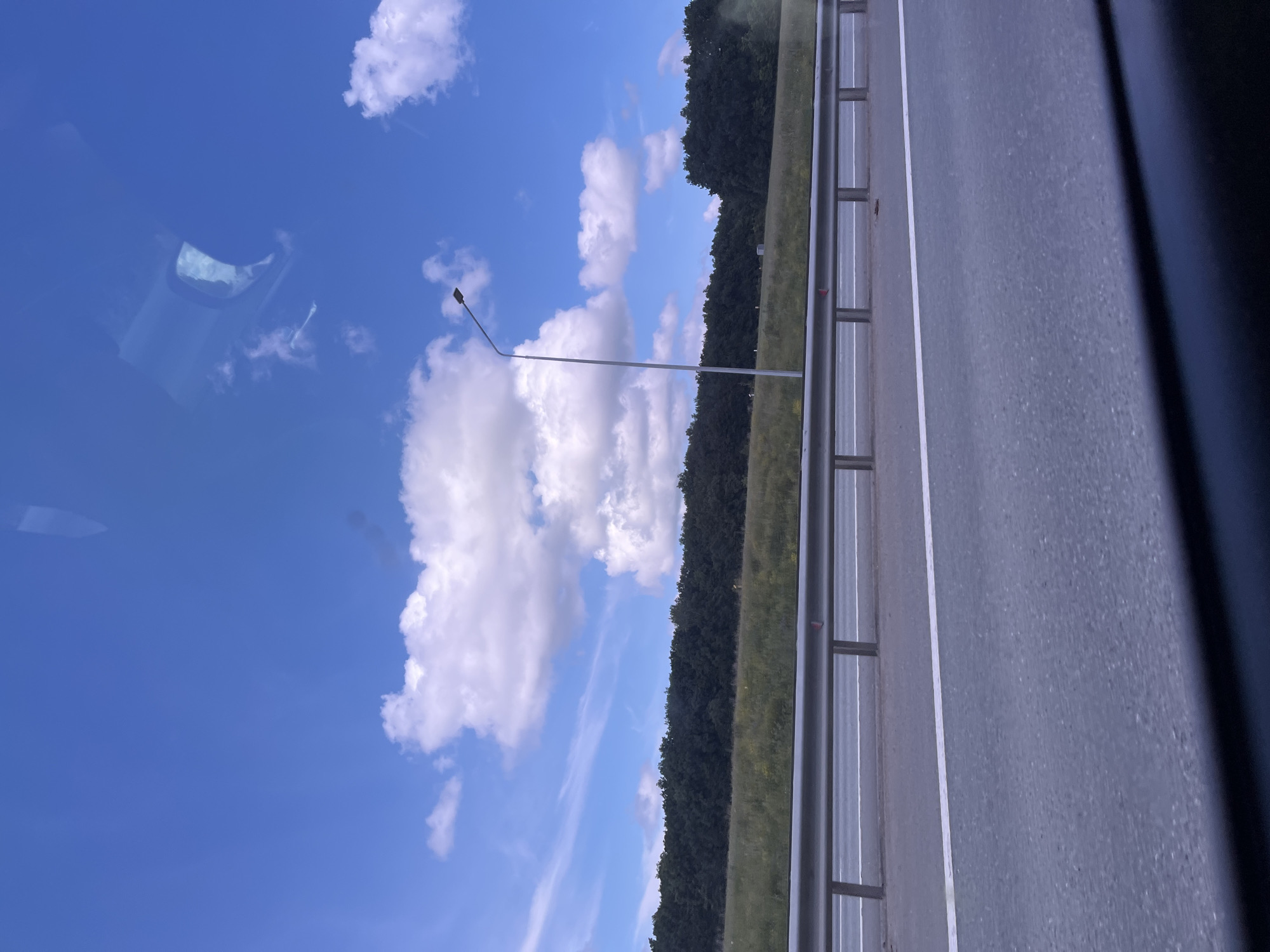 The image depicts a roadside view with a highway, guardrail, grassy median, and a line of trees under a bright blue sky with scattered white clouds. A lone street lamp stands on the median. The scene is devoid of people or any active events. The location is identified as being in the city of Bol'shoe Aksakovo, Russia. The perspective suggests the photo was taken from within a moving vehicle.