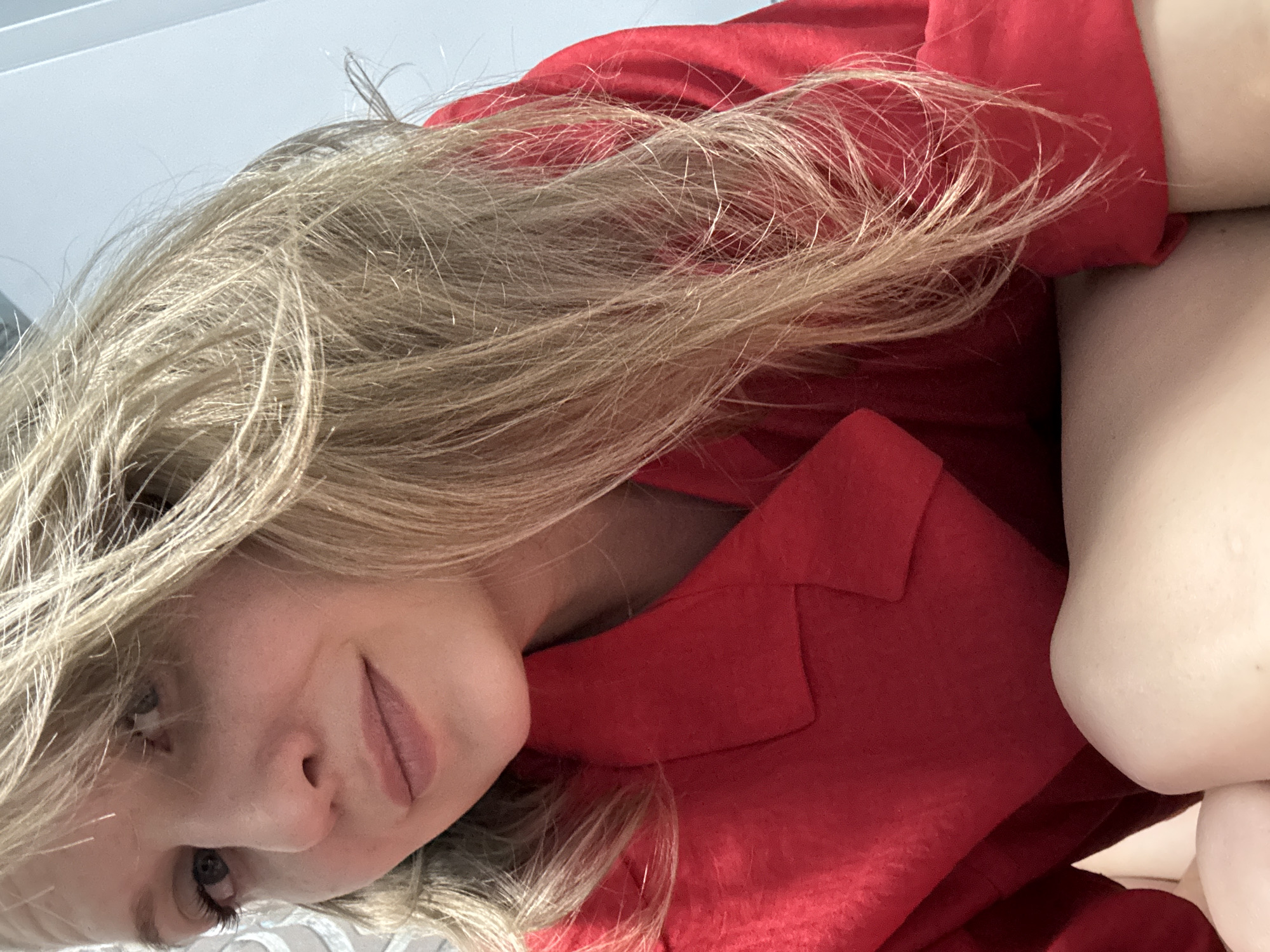 A woman with blonde hair wearing a red garment is depicted at an angle. Her face is visible, with a subtle smile. A pale arm is also partially visible. The location is not discernable from the image.