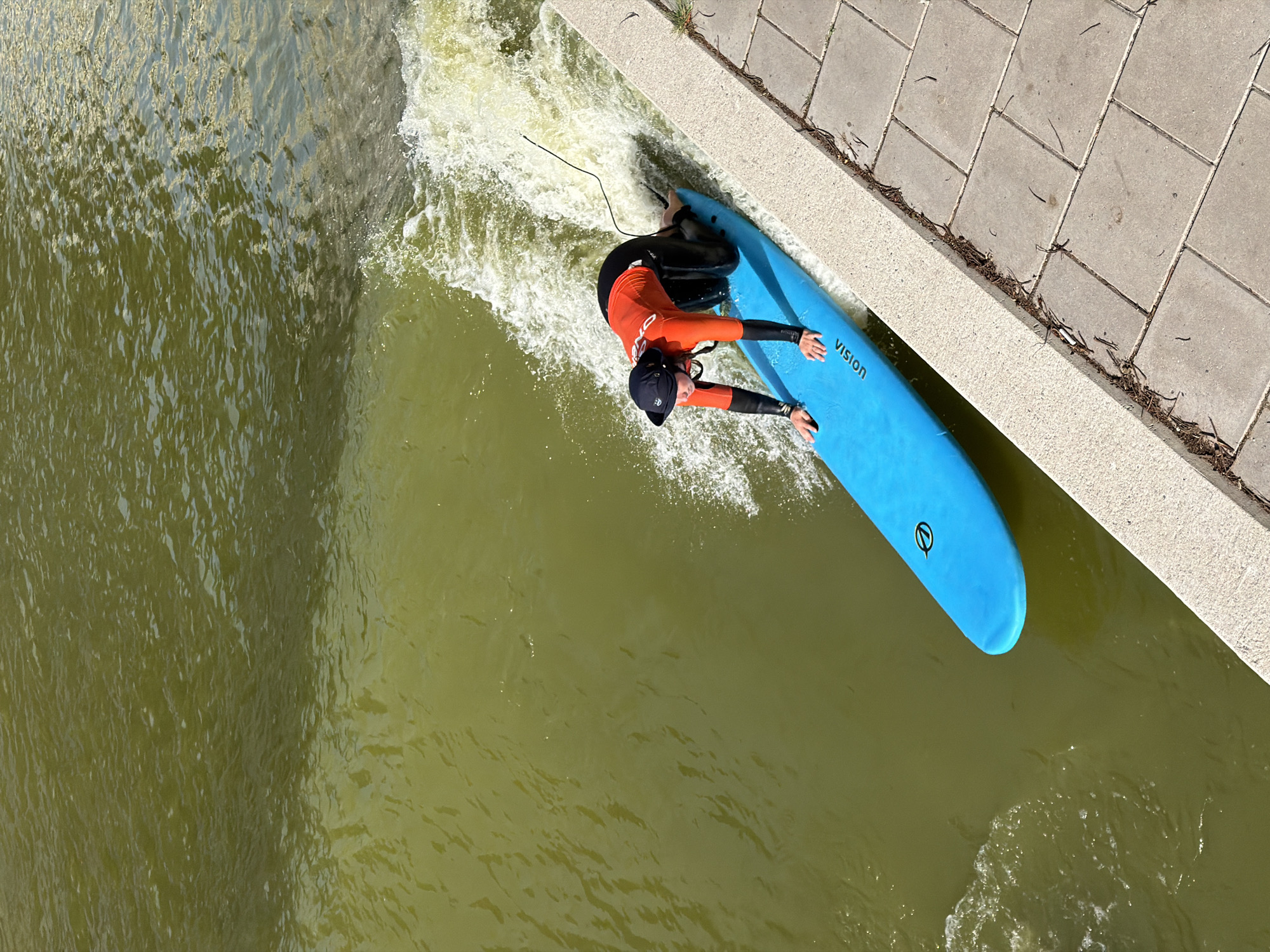 A person in a wetsuit and swim cap is surfing on a blue surfboard in a river. The river is green and has a white wave. The person is kneeling on the surfboard and holding onto the sides. The surfboard has the word "VISION" on it. In the background, there is a paved area with some dry leaves. This is likely the artificial wave at the Stokvisdijk in Rotterdam, Netherlands.