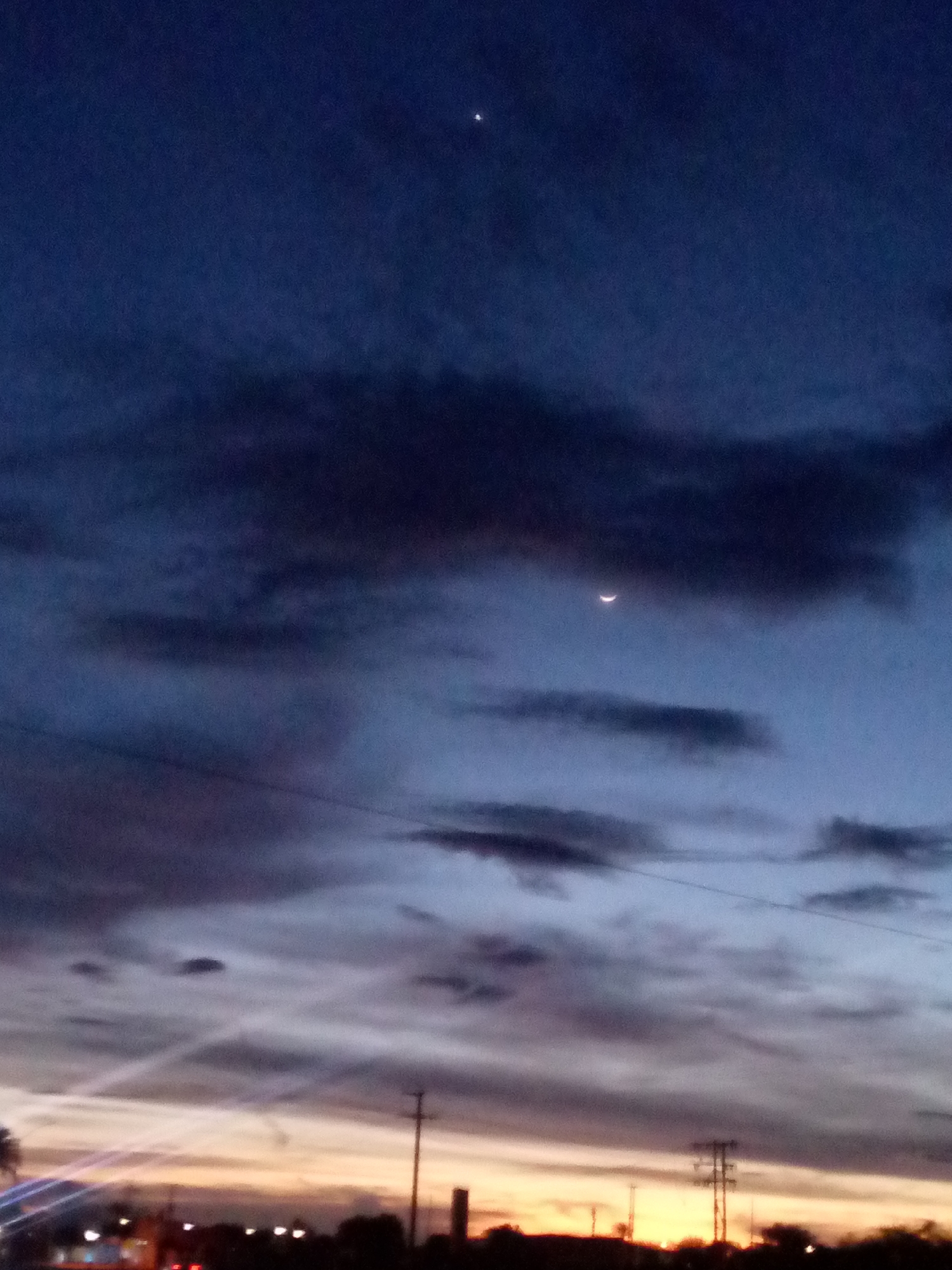 The image shows a twilight sky over a semi-urban landscape. In the upper portion of the sky, two celestial bodies are visible: a bright star or planet on the left and a crescent moon on the right. The sky is a gradient of deep blue at the top transitioning to lighter blues and purples, with streaks of orange and yellow near the horizon. Several dark, wispy clouds are scattered across the sky. Below the horizon, silhouettes of trees and structures, including utility poles and what appear to be buildings with lights, are visible. The setting suggests an evening scene in a populated area. The location is specified as Las Guevaras, Venezuela, indicating a likely terrestrial view from that region during twilight hours.