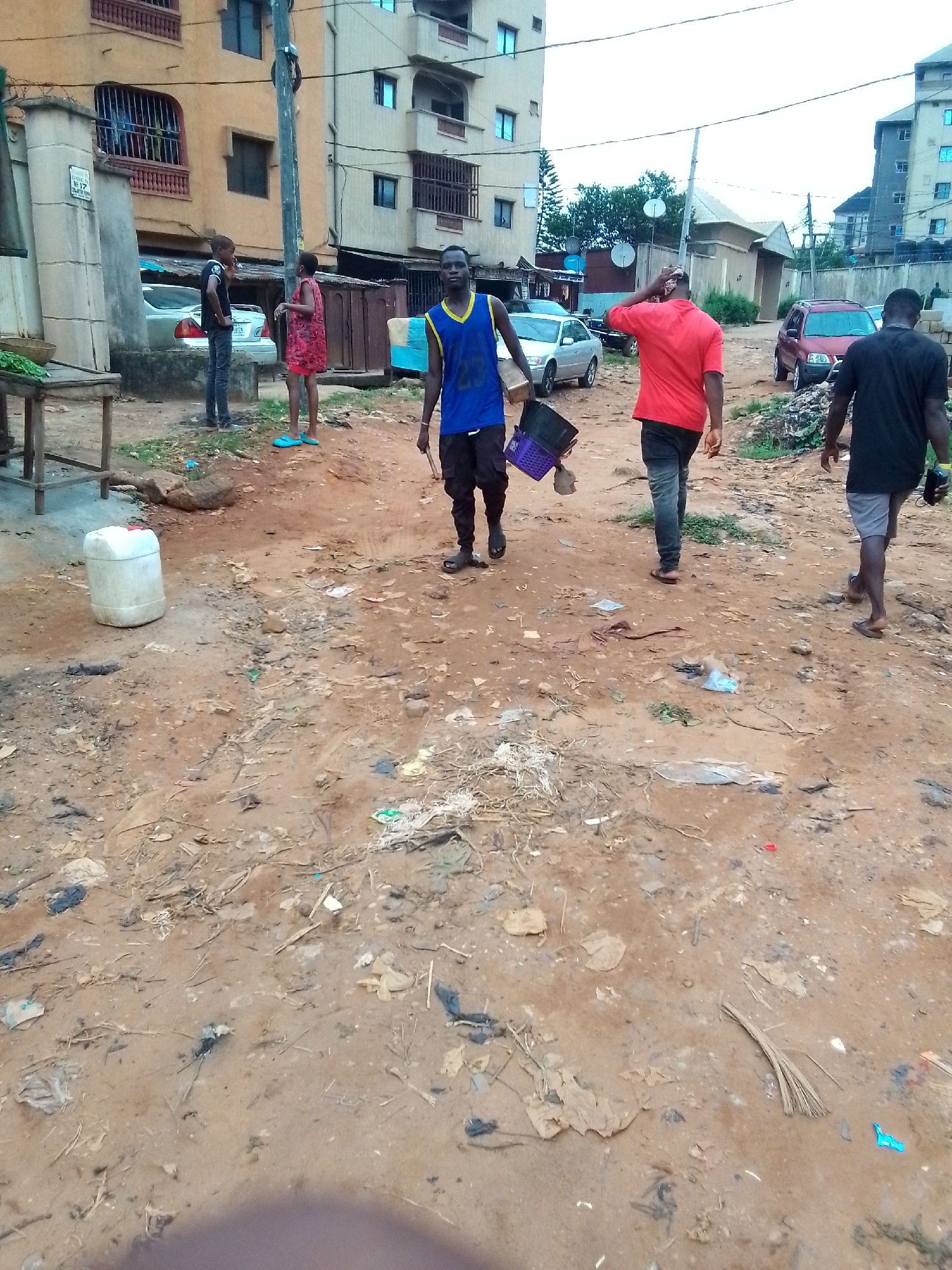 The image captures a bustling street scene in Obosi, Nigeria, under an overcast sky during daytime. The unpaved ground is reddish-brown dirt, extensively littered with various debris including plastic bags, wrappers, and organic refuse, suggesting a lack of formal waste disposal.
Multiple individuals populate the scene, engaged in daily routines. In the foreground and mid-ground, three men are walking away from the viewer. The nearest man, dressed in a blue and yellow sleeveless top, carries purple and black containers. Ahead of him, a man in a red t-shirt walks with his hand touching his head, while another man in a black t-shirt and grey shorts walks further to the right.
On the left side of the frame, near a multi-story building, a young man in a black t-shirt and jeans stands facing a young woman or girl in a red patterned dress and blue sandals. Behind them, a silver sedan is parked. Another man stands further back on the left, near a wooden table or stall where a white plastic jerrycan rests on the ground.
The background is dominated by multi-story residential buildings made of concrete or stucco, featuring barred windows and balconies. A prominent utility pole stands in the mid-ground. Other vehicles, including a red SUV, are visible further down the road. Satellite dishes are mounted on several buildings.
Visible text "LOCS No 17" is faintly discernible on a pillar or wall on the left side of the image, likely indicating a property number. The overall impression is of an active, lived-in urban environment characteristic of a busy residential area.