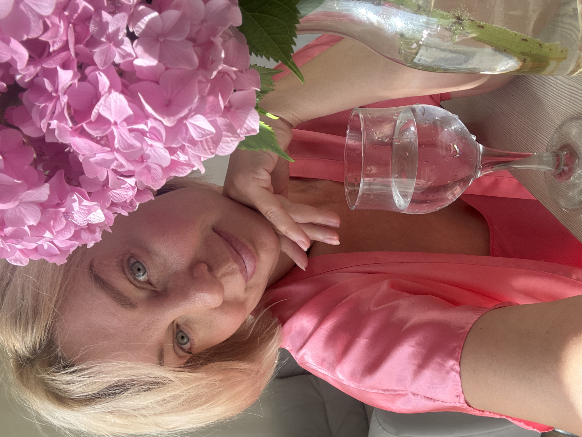 A woman with short blonde hair and light blue eyes is pictured in a close-up selfie. She is wearing a pink satin robe and her left hand is resting on her chin. To her right, a cluster of pink hydrangeas is visible, and in the foreground, a wine glass filled with water and a bottle with a green stem are placed on a wooden table. The background appears to be an indoor setting with a hint of natural light. The scene does not provide any information or context related to Sochi, Russia.