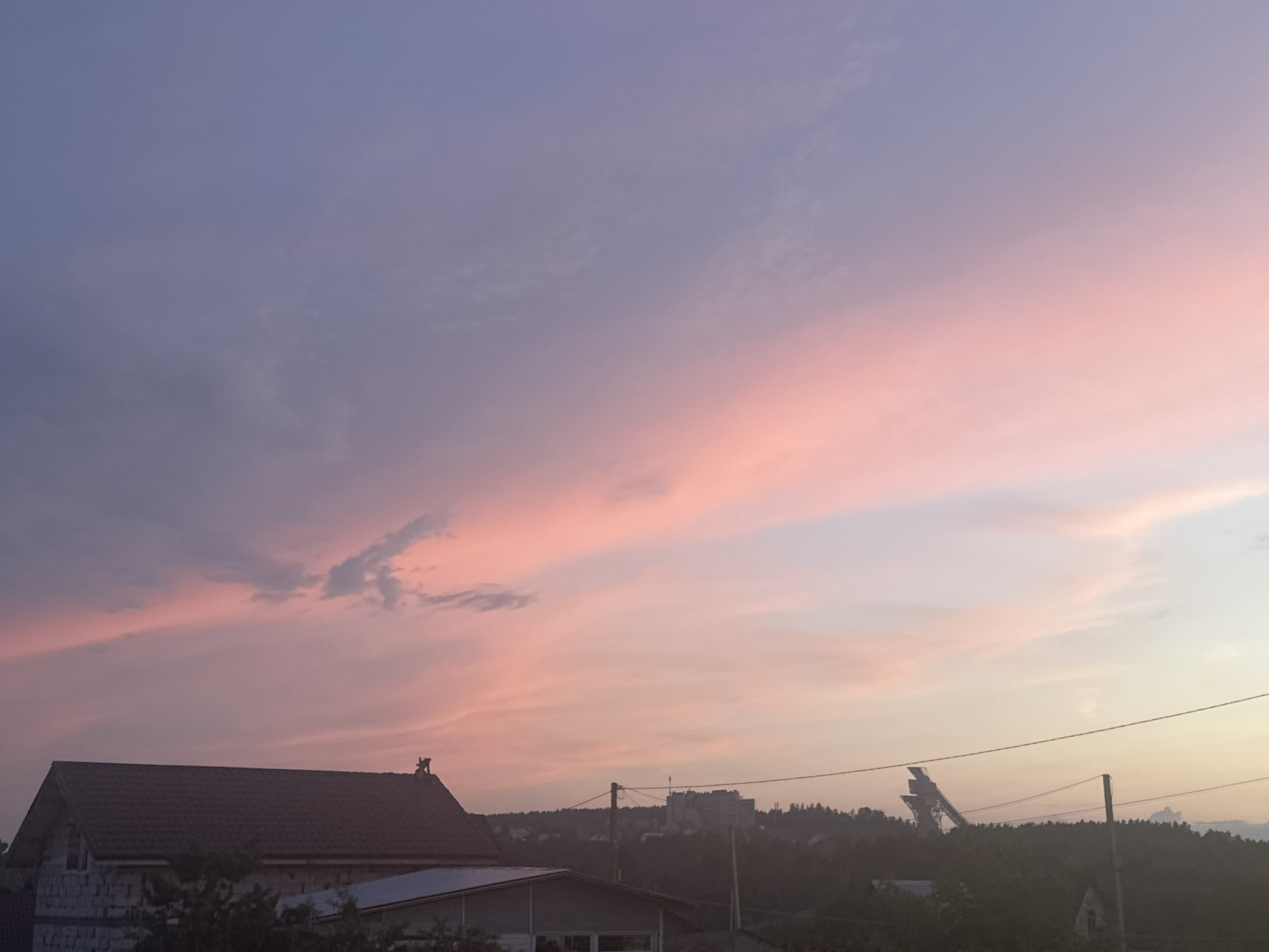 This is a medium-angle shot of a dusky sky over a rural or suburban area, likely near Прикамский, Russia. The sky is painted with soft hues of purple and pink from a setting sun. In the foreground, a dark-roofed house with white brick walls is visible on the left. Further into the scene, a distant row of trees separates the houses from a more developed area with buildings and a prominent ski jump structure. Power lines crisscross the sky and obscure parts of the landscape. The overall impression is one of quiet twilight.