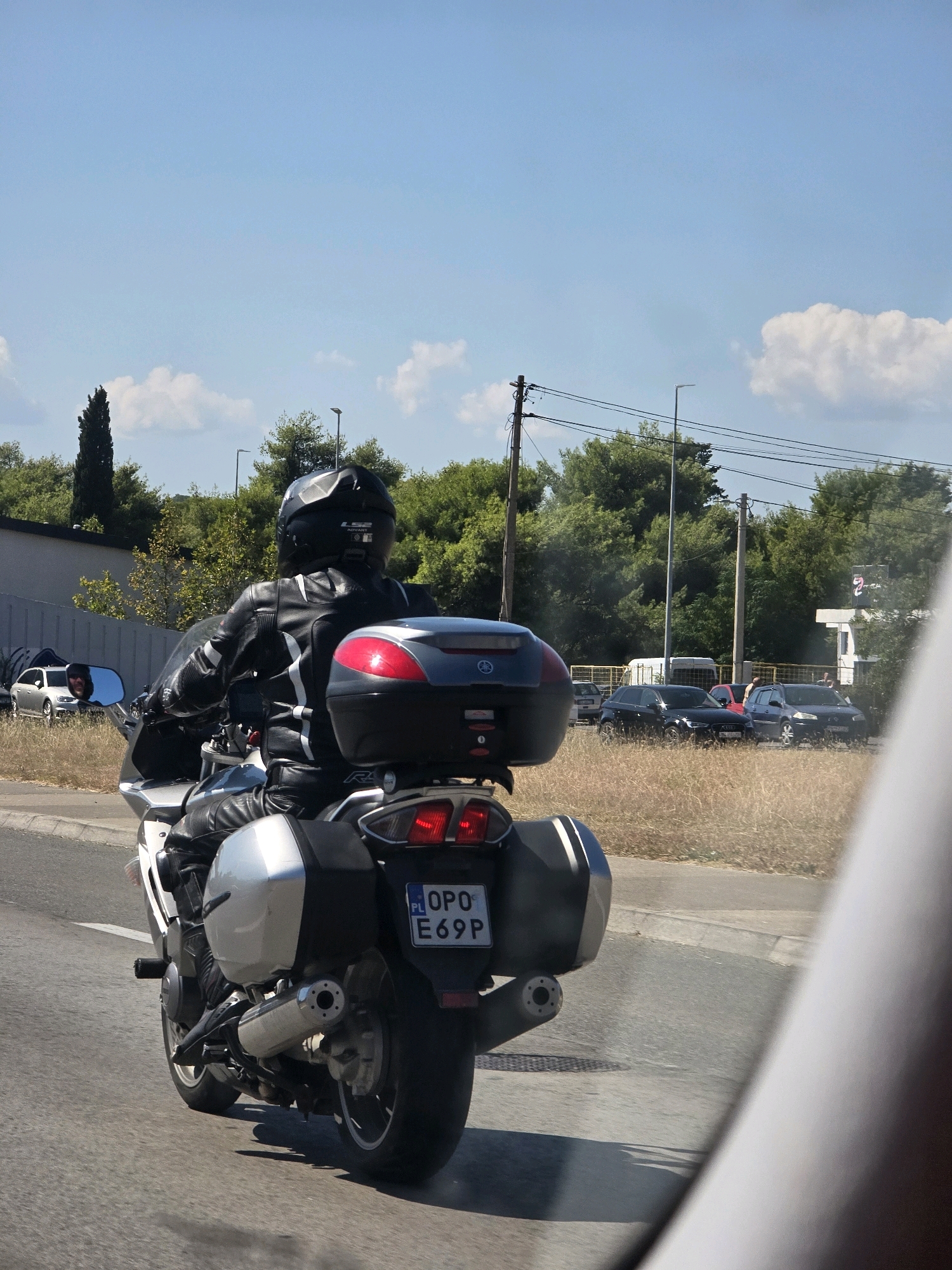 A person wearing a black motorcycle helmet and black leather motorcycle suit rides a silver and black motorcycle on a paved road. The motorcycle has a silver top case with red accents and two silver saddlebags on the sides. The license plate on the back of the motorcycle reads "OPO E69P". The rider is looking ahead, and the motorcycle is in motion, creating a slight blur on the road. In the background, there are trees, a street light, and several cars parked on the side of the road. The sky is blue with some white clouds. The scene appears to be in an urban or suburban area, possibly on a sunny day. The foliage suggests a temperate climate. Based on the license plate format, the location is likely in Montenegro, and the style of the vehicles and surroundings are consistent with Podgorica.