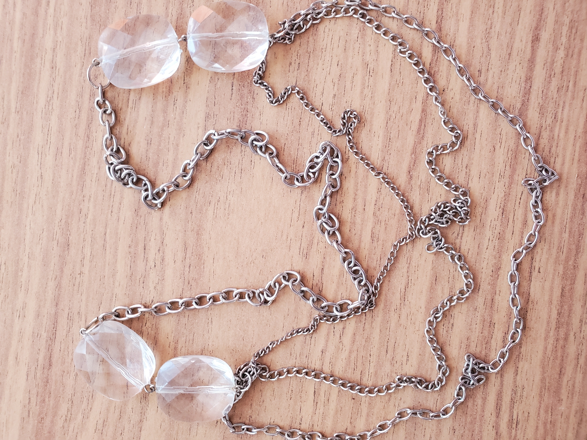 A silver-toned chain necklace with four clear, faceted oval beads is laid out on a wooden surface. The beads are of varying sizes, with the largest being at the top left and bottom right of the frame, and smaller ones positioned to their left and right. The chain is comprised of two distinct links, a thicker oval link and a thinner, more intricate loop. The wooden surface displays a natural grain pattern and appears to be a tabletop. There are no people or other objects visible in the image. The scene is static and the lighting is even, suggesting an indoor setting. There is no information provided that allows for the determination of the location in Tiraspol, Moldova.