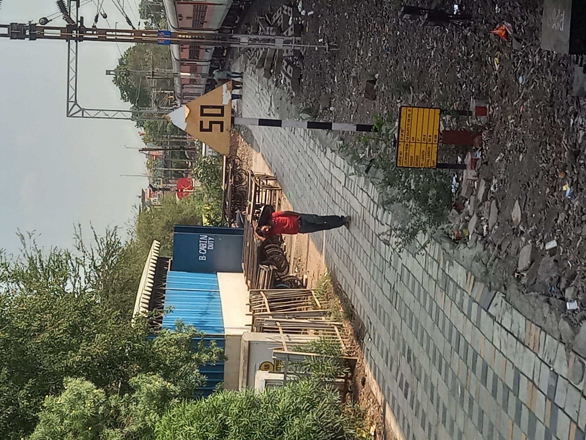 The image depicts a railway scene likely in Bhopal, India. A man in a red shirt and dark pants walks along a paved walkway. To the left, there's a blue structure labeled "B. CABIN DUTY," partially obscured by trees. Railway tracks and a train are visible in the background, along with overhead power lines. A triangular yellow sign with "50" is displayed, next to a sign with a yellow table on it. Debris and gravel litter the ground beside the walkway.