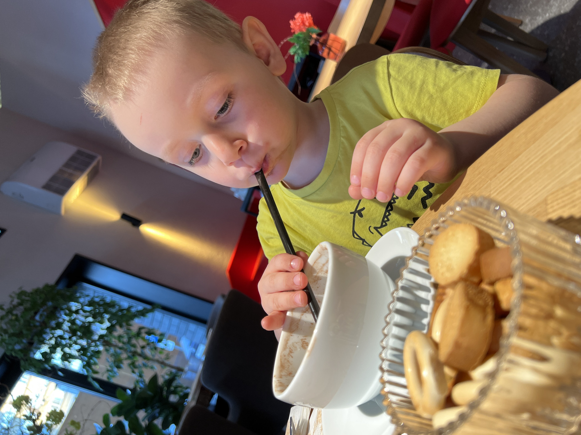 A young boy with blond hair, wearing a yellow t-shirt with a black dinosaur print, is drinking from a white mug with a black straw at a table. A clear glass bowl filled with small, round crackers or bread rings sits next to the mug. The background features a window with plants, a dark chair, a red wall, and a red floral arrangement on a shelf. The image appears to be taken indoors, possibly in a cafe or restaurant. While it’s impossible to confirm the location with certainty from just the image, based on the tags, it is possibly in Mytishchi, Russia.