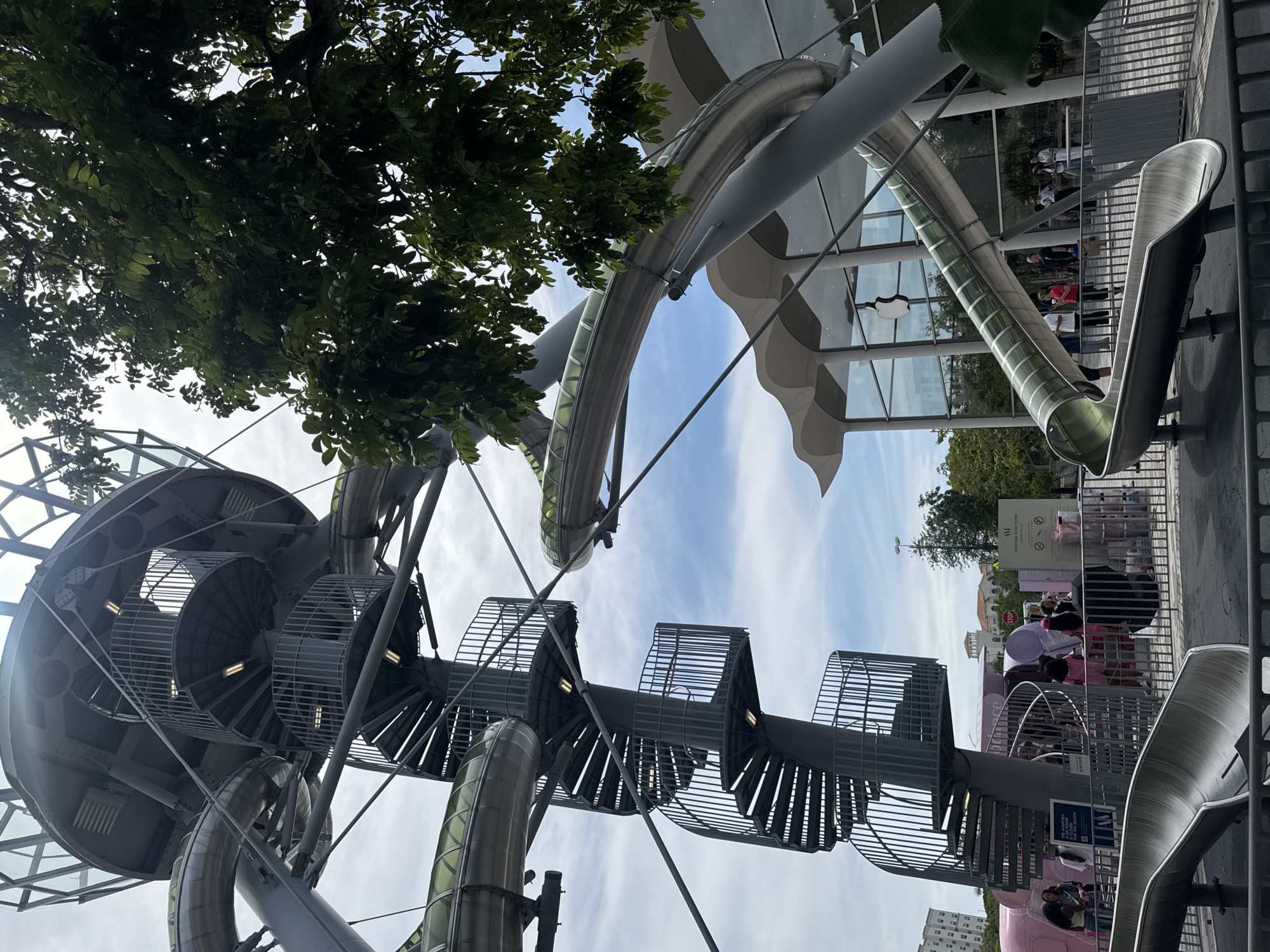 This is a low-angle shot of a large, modern outdoor structure featuring spiral staircases and winding, enclosed metal slides. The structure is gray and metallic, with some parts of the slides having green translucent coverings. The background shows a building with a distinctive wavy roofline and large glass windows. An Apple logo is visible on one of the windows, indicating this is likely the Apple Store Aventura, located in Aventura, Florida, United States of America. Lush green trees with dense foliage are on the right side of the frame, partially obscuring the sky, which is partly cloudy and blue. In the foreground, there are metal barriers and the bottom ends of the slides. A few people are visible in the mid-ground, some standing near informational signs, and others in the background near the building. The scene appears to be a public space or a recreational area adjacent to the retail building.