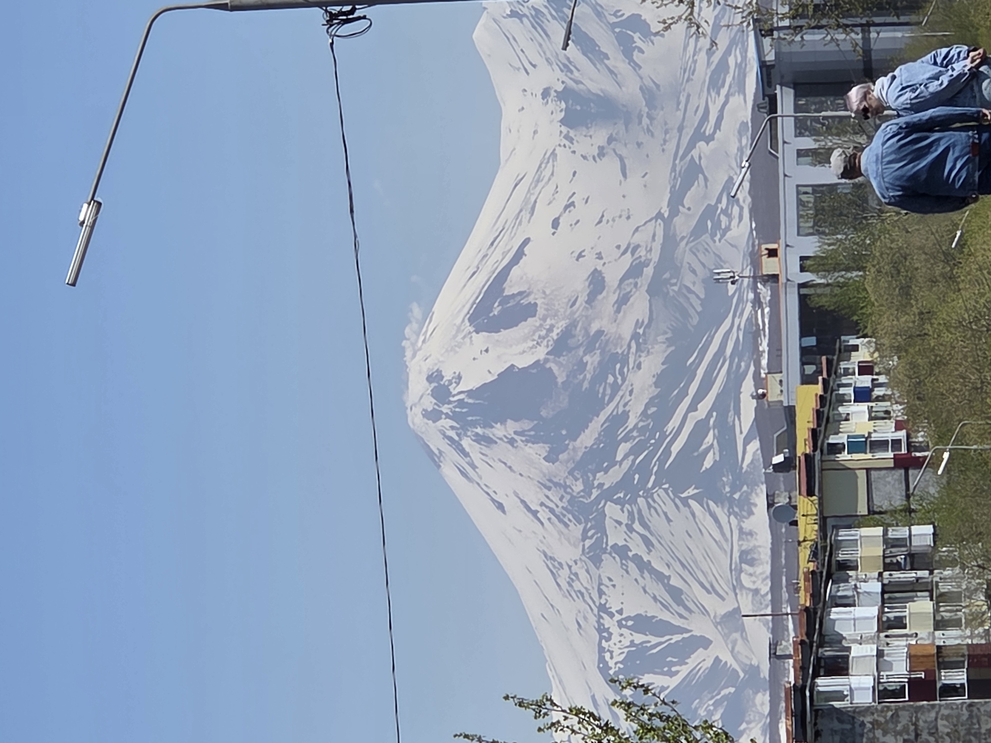 The image captures a vast snow-capped mountain range under a clear blue sky, likely Mount Ararat, dominating the background. A prominent peak, possibly a volcano, shows a slight atmospheric haze or cloud near its summit. In the midground, a diverse urban landscape unfolds, featuring a row of weathered, multi-story residential buildings with various colored balconies and facades on the left, and more modern, light-colored buildings with reflective windows on the right. A satellite dish is visible on the roof of one older building, and a cellular tower stands on a modern building's roof. Green foliage from trees is interspersed throughout the midground. A modern street light, with an elongated silver fixture, is prominently positioned on the right, extending into the upper portion of the frame. Dark power lines or cables stretch horizontally across the image, partially obscuring the mountain. In the lower right foreground, two individuals are seen from behind, walking away from the viewer. One wears a blue denim jacket and a light cap, while the other wears a light blue denim jacket and has short, light-colored hair. The scene is located in the city of Unknown, Unknown.