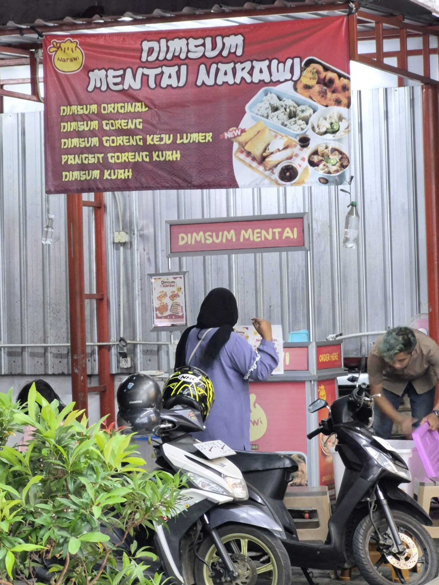 The image depicts an outdoor food stall in Purworejo, Indonesia, specializing in dim sum. A large red banner prominently displays the establishment's name, "DIMSUM MENTAI NARAWi," alongside images of various dim sum dishes. Below the main banner, a smaller sign reads "DIMSUM MENTAI."
In the center of the scene, a person wearing a dark hijab and a purple top is standing behind the counter, appearing to interact with a customer or prepare an order. To the right, another person with green-tinged hair, wearing a brown shirt and blue jeans, is leaning over and appears to be handling a pink plastic container. Two motorcycles are parked in front of the stall, one black and one white, with helmets placed on them. Lush green foliage is visible in the foreground, partially obscuring the motorcycles and stall. The stall itself has a corrugated metal background and a wooden counter. The environment suggests a warm, possibly humid, day, typical of the region.