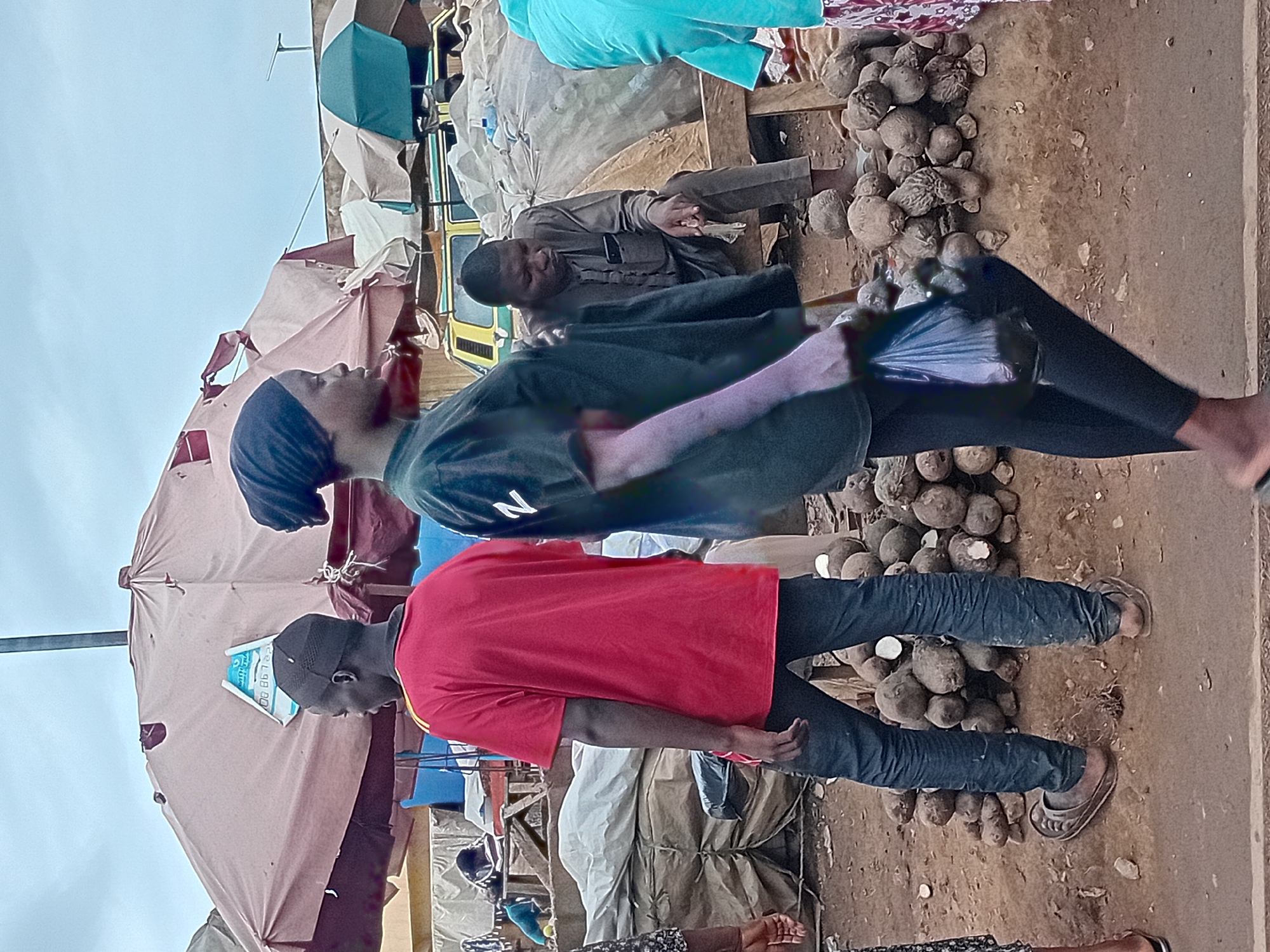 In Maiburiji, Nigeria, an outdoor market scene unfolds under an overcast sky. The foreground is dominated by a woman in a black headscarf and black leggings, walking from right to left, carrying a black plastic bag. She is positioned in front of a pile of yams. To her left, a man wearing a red t-shirt and dark pants stands facing away from the camera, looking towards a large, tattered beige umbrella. Behind the man in red, another man is seated on a wooden bench, wearing a grey traditional outfit and holding a cigarette. The background features various market stalls with umbrellas, a yellow vehicle, and more piles of produce. The ground is dusty and uneven, typical of a market environment.