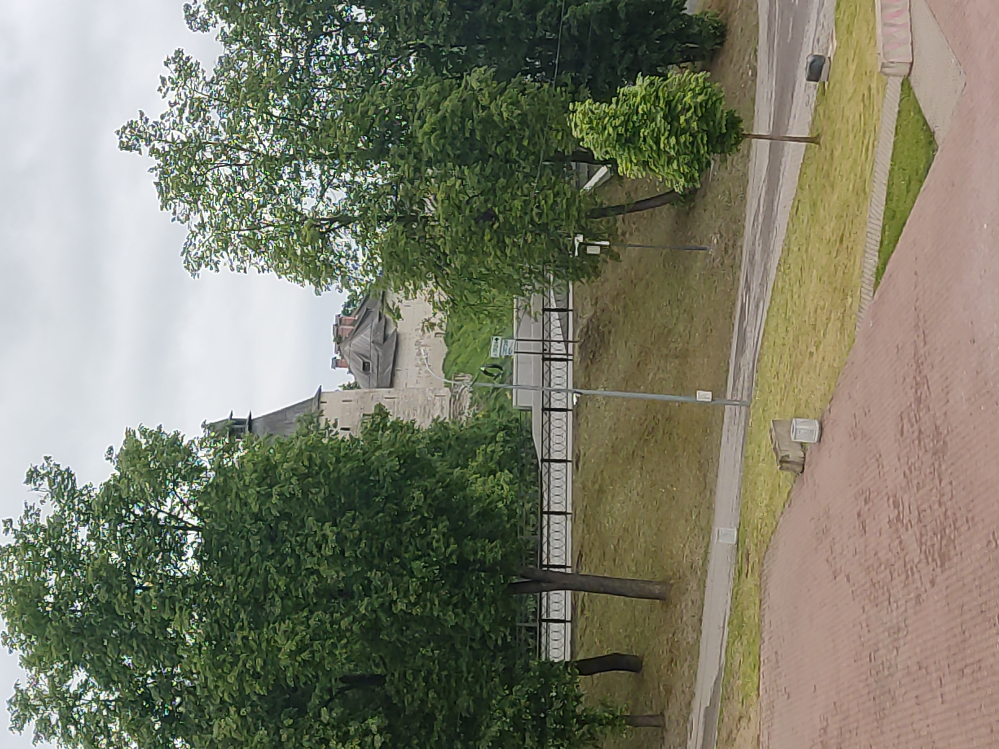 This image shows the Pskov Kremlin in Pskov, Russia, viewed from an elevated position. Trees with dense green foliage frame the view. A gray stone wall and structures with pitched roofs are visible in the background. A road runs along the base of the Kremlin walls, bordered by a metal fence. Streetlights are positioned along the road. The foreground features a brick-paved area.