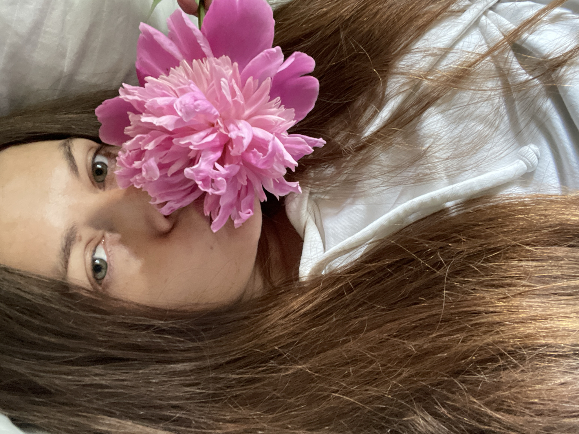 A medium shot shows a woman with long, brown hair and a white hoodie holding a large, pink peony to her face, obscuring her mouth. Her left eye is visible, looking towards the camera. The background is a white, textured surface, possibly a bedsheet. The image is a selfie, taken from a slightly elevated angle. The location is not specified in the image or its context.