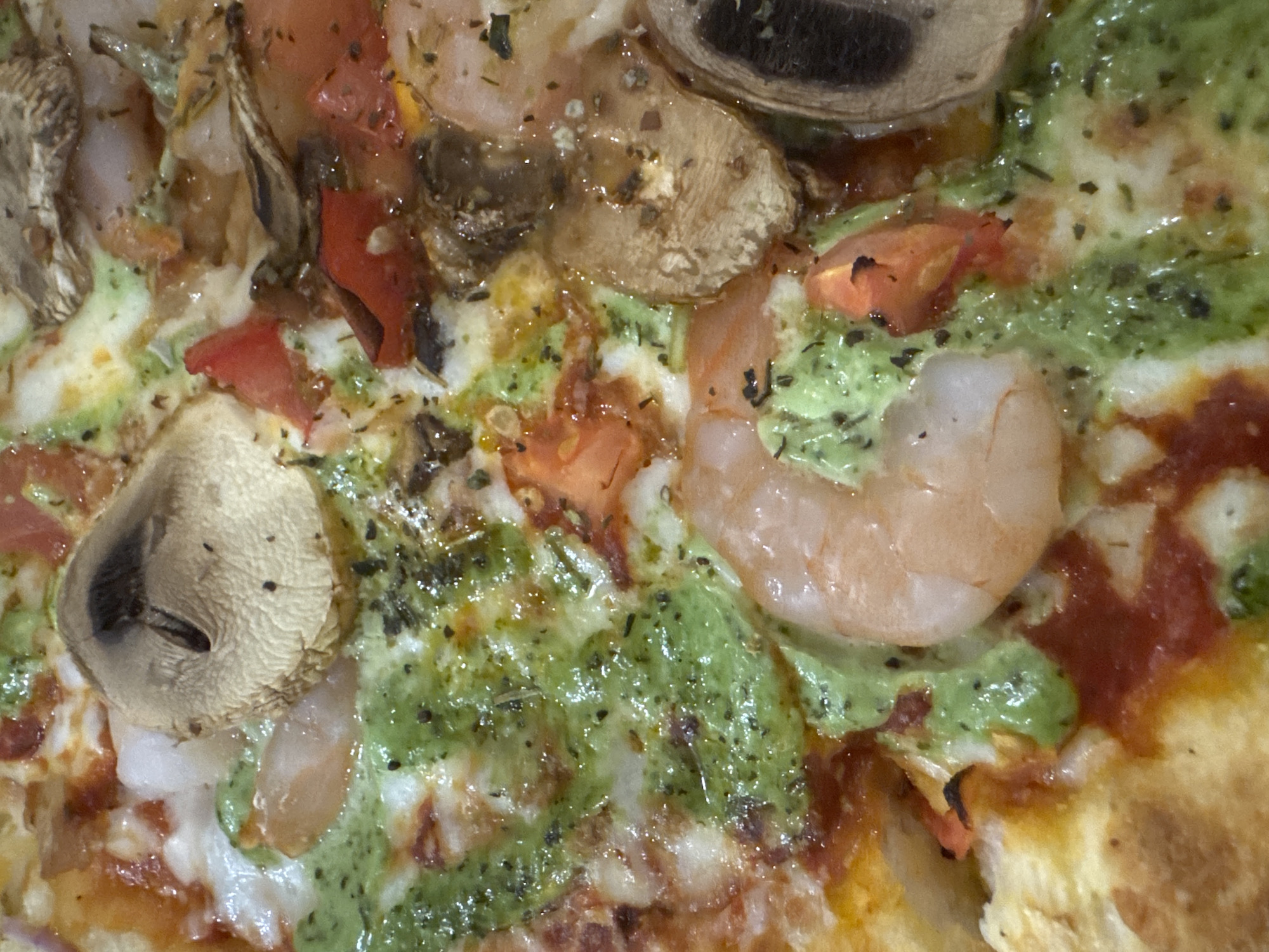 The scene, located in Votkinsk, Russia, is a forensic close-up, top-down depiction of a pizza section. The surface is densely covered with cooked, curled pinkish-orange shrimp, slices of brown mushrooms, and diced red tomatoes. Melted white and light-yellow cheese forms a patchy layer over a visible red tomato sauce base. A vibrant green, creamy sauce is generously drizzled over the entire surface, accentuated by scattered dark, dried herbs. Portions of the golden-brown pizza crust are visible along the edges and bottom of the frame.