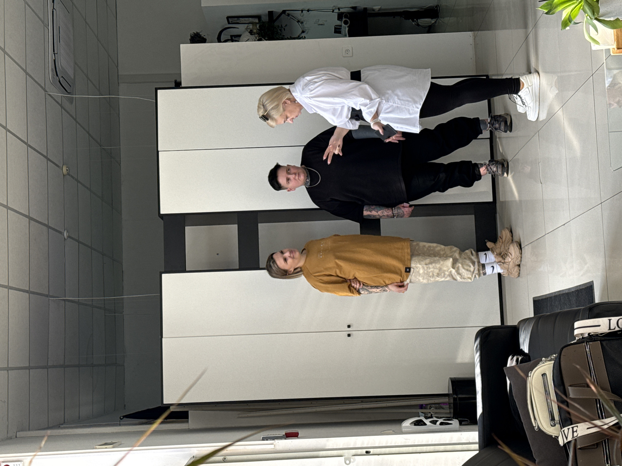 This image, captured indoors in Rostov-on-Don, Russia, depicts three adult females engaged in a conversation within a modern, brightly lit commercial or office space.
The environment features a white tiled floor reflecting ambient light, a white modular ceiling with visible grid lines, an overhead air conditioning unit, and two ceiling-mounted smoke detectors. The background consists primarily of large, white modular cabinets or room dividers with black handles. A strong light source from the left casts distinct shadows and reflections. In the foreground, to the left, a black sofa with dark pillows holds a light-colored purse and a larger dark bag with a white strap. A small green plant is partially visible in the bottom right corner.
From left to right:
1. An individual with short, light brown hair and visible tattoos on both arms is positioned on the left, facing right. She wears a loose mustard-yellow long-sleeve t-shirt, light-colored patterned cargo pants, white socks with a black logo, and chunky light brown athletic shoes. Her expression is a slight smile as she looks towards the two individuals on the right.
2. A second individual, positioned center-right, has short dark hair and visible extensive tattoos on both arms and neck. She wears a black t-shirt, black pants, a silver chain necklace, and dark athletic shoes. Her expression is neutral as she faces the third individual. She holds a dark object, possibly a mobile phone, in her left hand.
3. A third individual, on the far right, has short blond hair and sunglasses resting on her head. She wears a long-sleeved white button-down shirt belted with a wide black belt, black leggings, and white platform sneakers. Her right hand is raised and open, fingers slightly spread, indicating active gesticulation during speech, directed towards the second individual. Her left hand is down and slightly open.