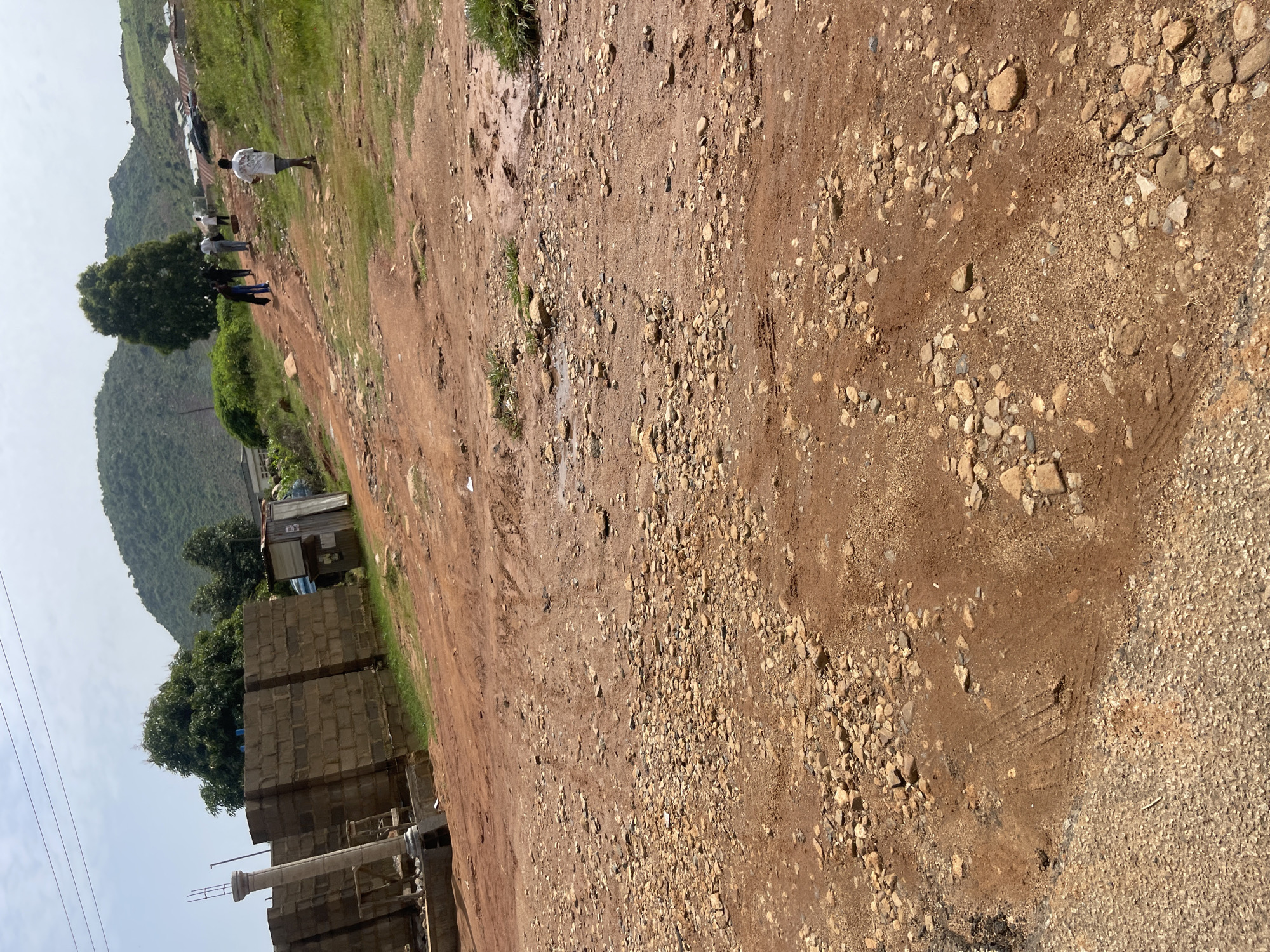 The image depicts a dirt road in Gwong, Nigeria, leading uphill towards a cluster of homes. The road is uneven and strewn with rocks and pebbles, with tire tracks visible in some sections. To the left, a partially constructed building with cinder blocks and a concrete pillar is apparent. A small wooden structure, possibly a kiosk, sits further up the road. Several people are walking up the hill. In the distance, a large tree stands prominently, with a lush green hillside and a hazy sky overhead.