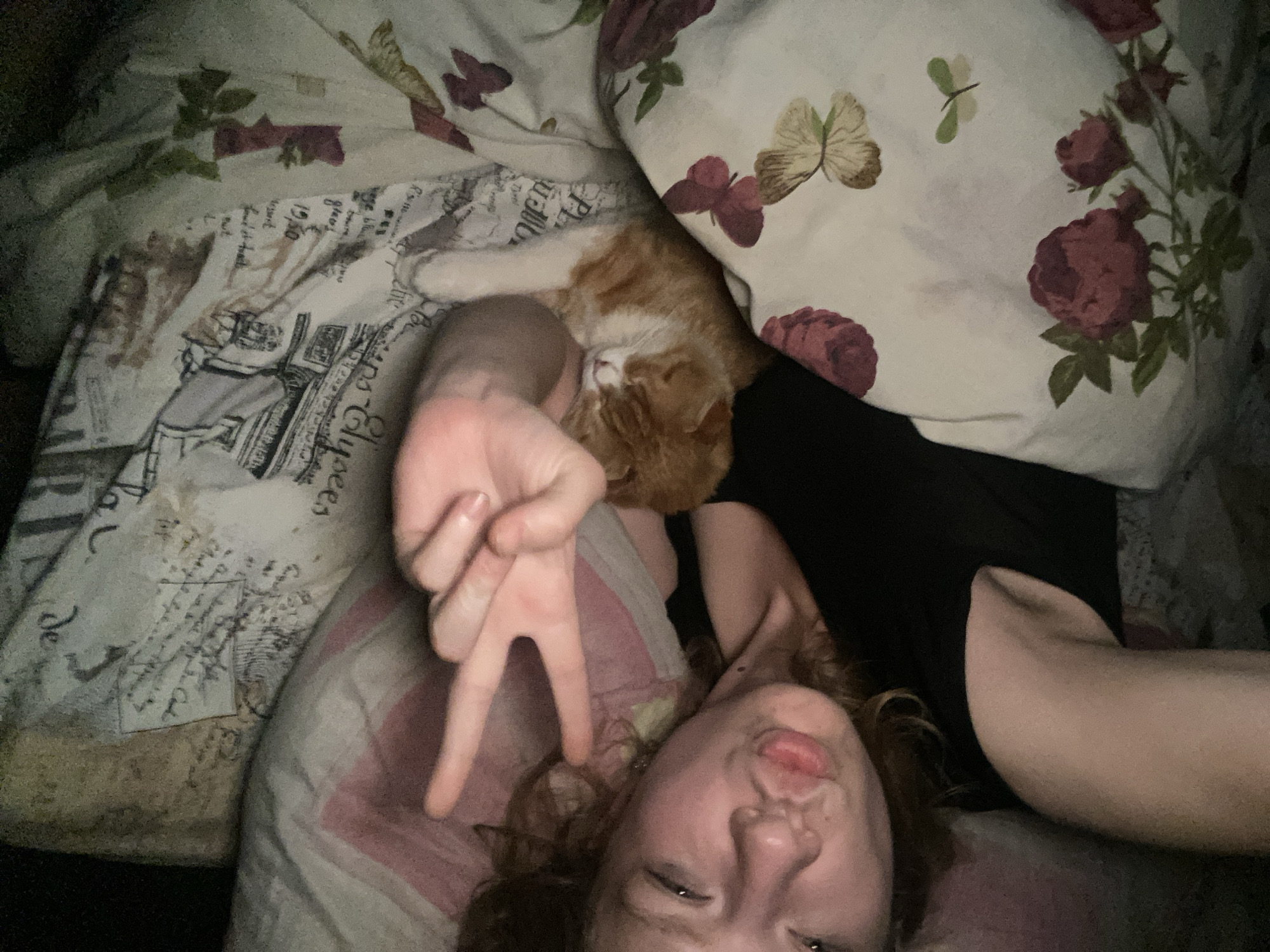 A person is lying in bed with an orange cat. The person is making a peace sign with their hand and sticking their tongue out. The bedding has a floral pattern on the pillow and an architectural pattern on the sheet. The location is not determinable.
