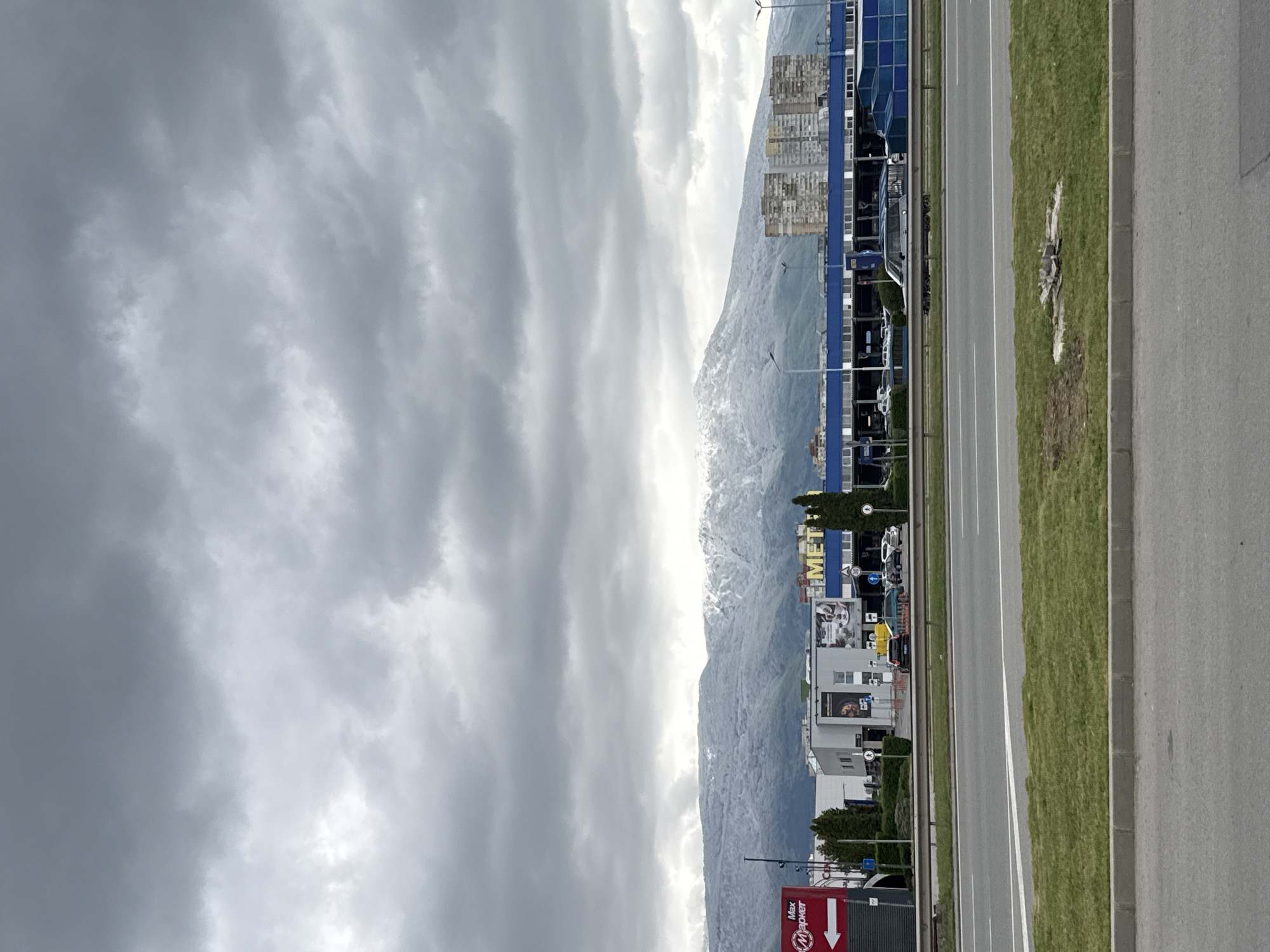 The image, when viewed horizontally, depicts an urban or suburban landscape dominated by a wide road, commercial buildings, and a majestic mountain range under an overcast sky.
In the foreground, a multi-lane asphalt road stretches horizontally, bordered by a grassy median strip and a concrete sidewalk or shoulder on the right. The road appears clear of traffic.
The midground is occupied by a series of commercial establishments lining the road. One prominent building features a blue facade with a large "METRO" sign, indicative of a Metro Cash & Carry store. Further down, a red sign with a distinct arrow pointing downwards and the Cyrillic text "Max маркет" (Max Market) is visible. Other smaller buildings and parked vehicles can also be seen in this area.
The background is dominated by a substantial mountain range, likely the Vitosha Mountains, given the Sofia, Bulgaria context. The peaks and upper slopes of these mountains are partially covered in snow, suggesting either winter or early spring.
The sky is heavily overcast with dense, grey clouds, indicating a gloomy or cool daytime atmosphere. No individuals are visible in the scene. The image captures a commercial zone at the foothills of mountains on a cloudy day.