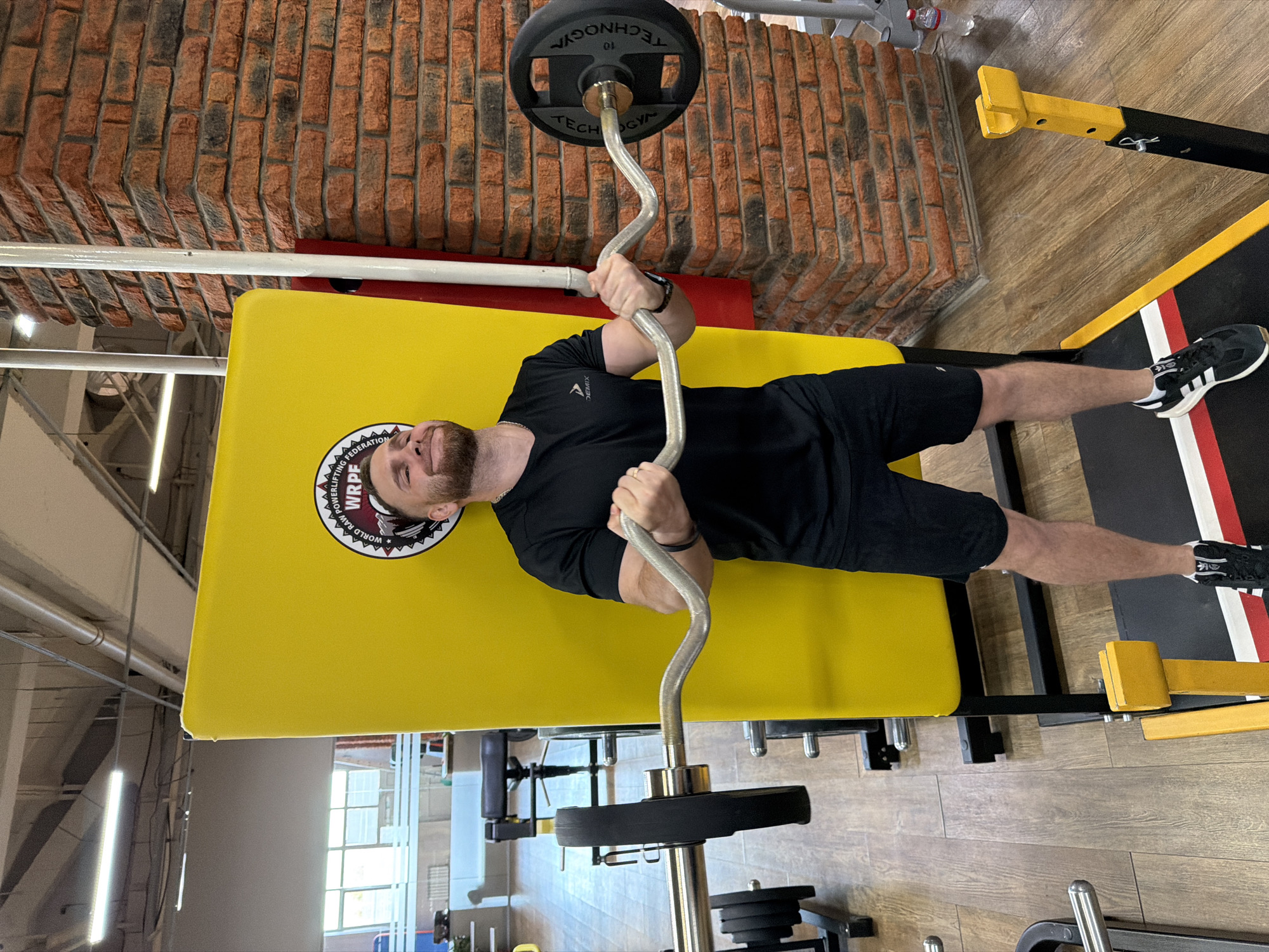 A man is performing a weightlifting exercise on a bench press in a gym. The bench is yellow and has a logo on it. The man is wearing a black t-shirt and black shorts. He is holding a curved barbell with weights on it. The gym has brick walls and a wooden floor. The location is in the city of Chaikovsky, Russia.