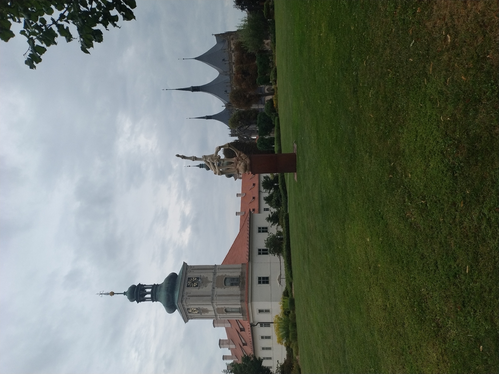 The image depicts a view of the Church of Saint Barbara in Kutná Hora, Czechia. In the foreground, a well-maintained green lawn slopes gently upwards towards a sculpture of a skeletal hand raised in the air. Behind the sculpture, the majestic Gothic spires of the Church of Saint Barbara dominate the skyline under a cloudy sky. To the left of the church, a section of the former Jesuit college, now the Gothic House, is visible, characterized by its red tiled roofs and a prominent clock tower with a green dome. Trees and bushes are scattered throughout the scene, adding to the park-like atmosphere. The overall lighting suggests an overcast day. There are no people visible in the image.