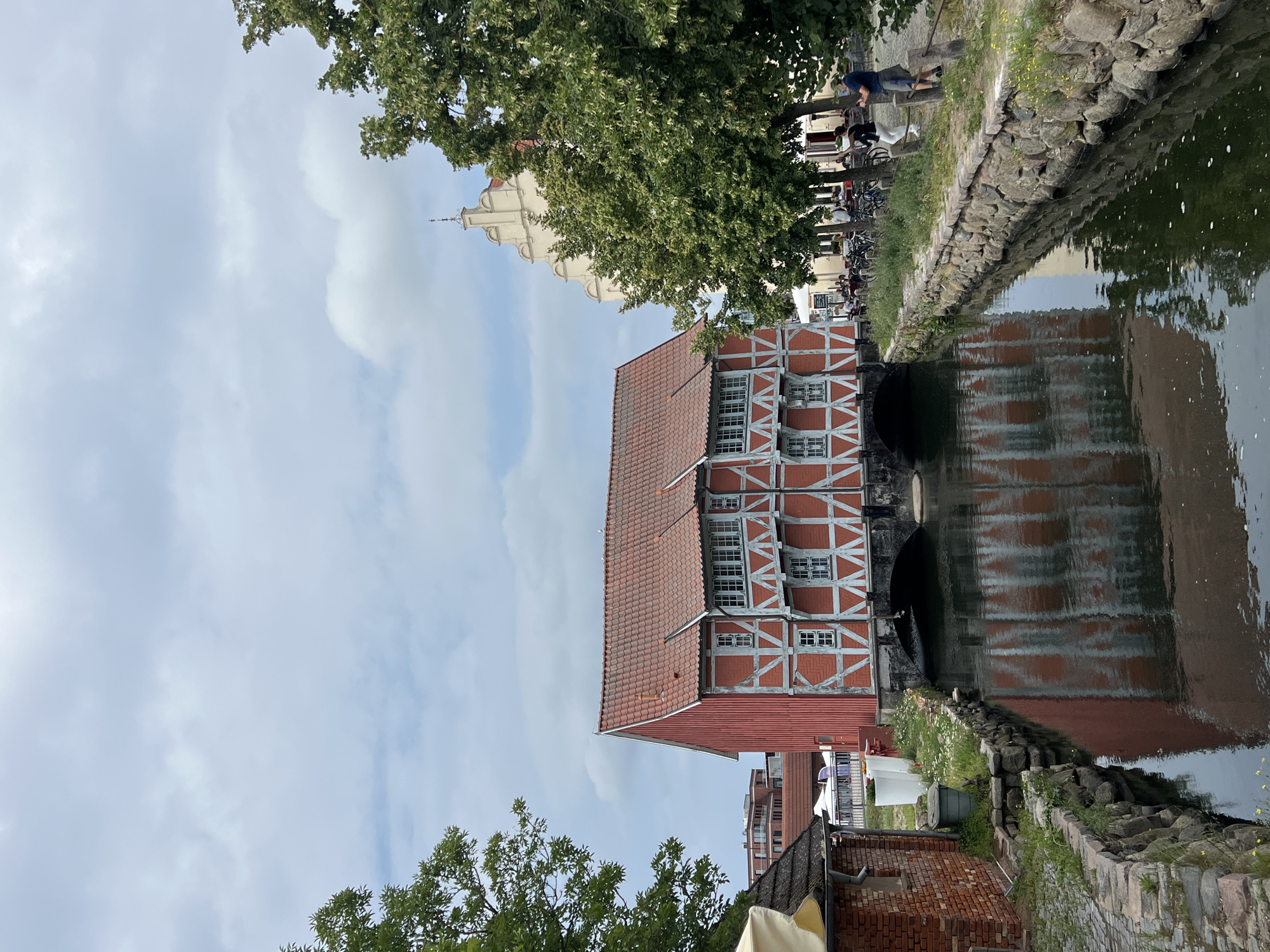 The image depicts a canal flowing through the historic town of Wismar, Germany. A distinctive red and white half-timbered building, which is a historic mill known as "Alter Speicher" or "Wassermühle," straddles the canal. The building has a red tiled roof and numerous white-framed windows. The canal's dark water reflects the building's facade.
On the right bank of the canal, a stone-lined embankment is visible, with grass and small yellow flowers growing along it. A paved walkway runs alongside the embankment. Several people are present on the right side. Two men are standing, with one appearing to take a photo with his phone. A woman in white pants and a dark top is also present, and a group of people are seated at tables outside a building in the background, suggesting a cafe or restaurant. Bicycles are parked near the walkway.
On the left bank, a portion of a red brick building with a dark roof is visible, with a yellow awning. A grey trash bin and some white planters are also present.
In the background, behind the mill, other buildings are visible, including a taller, light-colored building with a stepped gable. Trees with lush green foliage line the right bank of the canal, partially obscuring the background buildings. The sky is overcast with scattered clouds. The overall scene suggests a pleasant day in a picturesque European town.