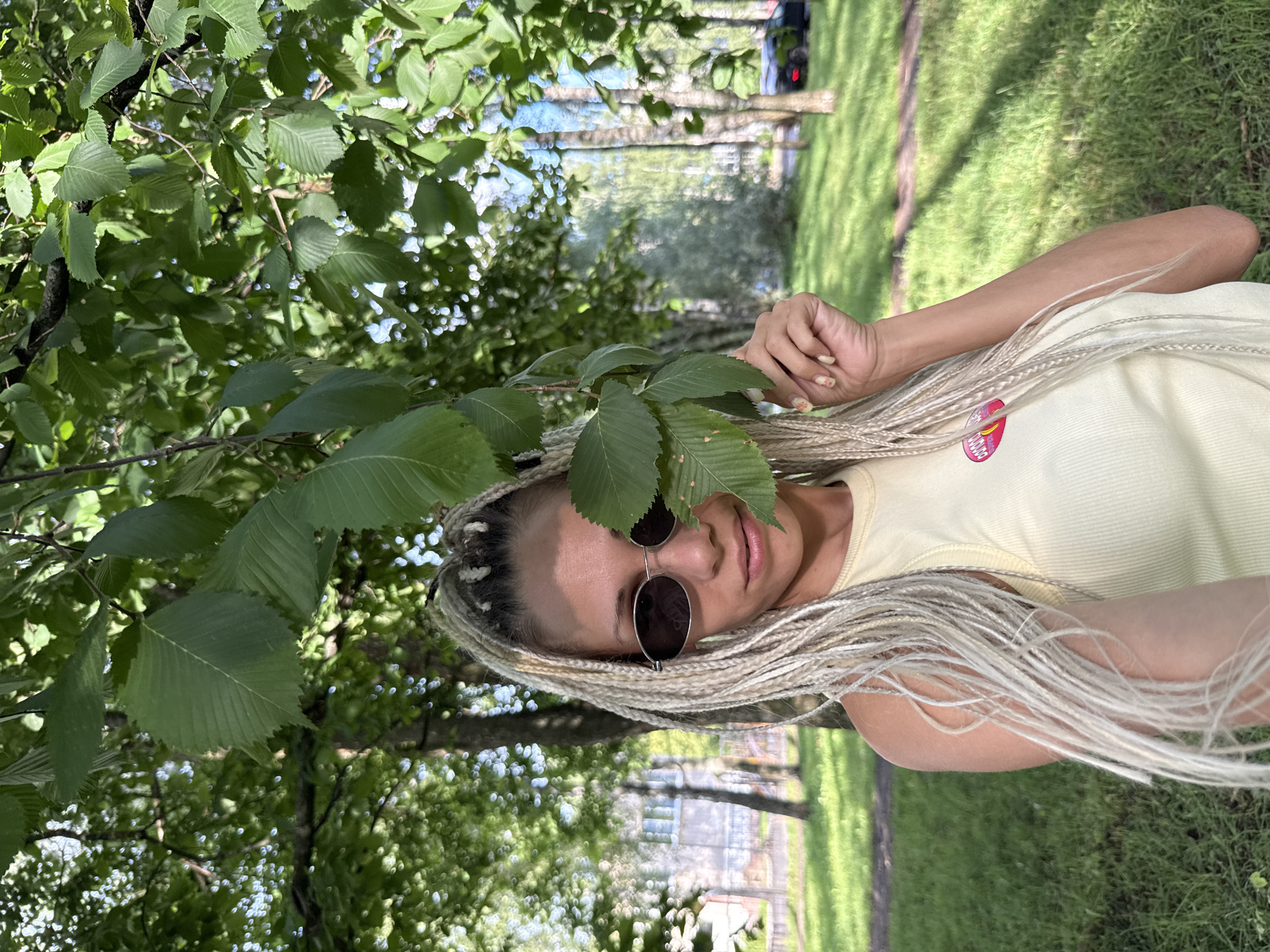 A woman with blonde braided hair and sunglasses is standing outdoors in a park in Yaroslavl, Russia. She is wearing a light yellow tank top with a circular graphic on the front. Green leaves from a tree partially obscure her face and arm. The background shows trees, a grassy area, and a building. Sunlight is filtering through the leaves, creating dappled lighting on the ground.