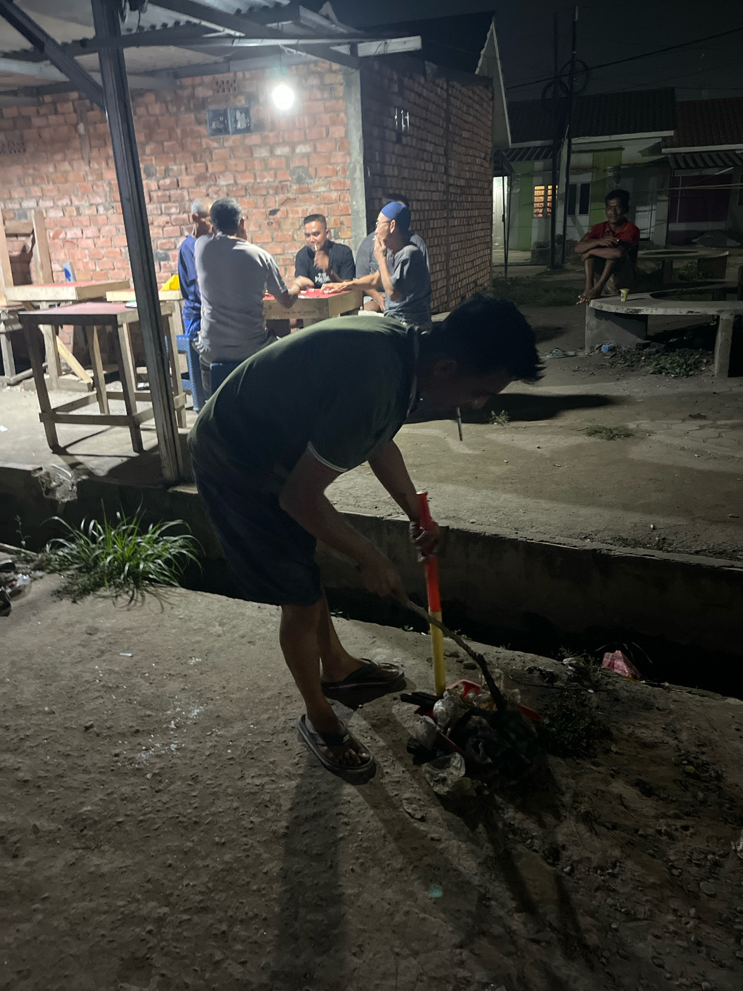 A man in a dark green t-shirt and dark shorts is depicted cleaning up trash with a stick in a low-angle, outdoor shot at night in Talang Kelapa, Indonesia. He is bent over, using the stick to gather debris that is accumulated in a gutter or drainage area. The trash includes plastic bags, wrappers, and other discarded items. He is wearing dark sandals. In the background, several other men are visible. Some are seated around a table under a makeshift roof structure constructed from corrugated metal and wooden beams, illuminated by a single bright light bulb. These men appear to be socializing or engaged in an activity like playing cards, with one man smoking. The environment suggests a casual, possibly informal gathering place, with brick walls and a concrete ground surface. Further in the background, residential structures with lit windows are dimly visible, indicating the urban setting. The overall scene conveys a sense of community life taking place after dark.