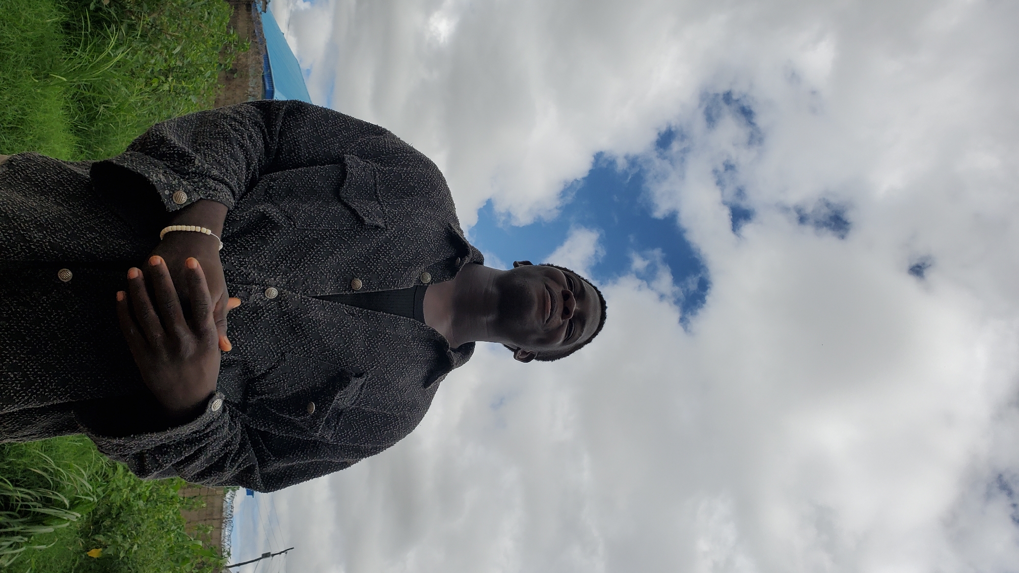 In Jalingo, Nigeria, a young Black man stands outdoors against a cloudy sky with a blue patch visible. He is wearing a dark, textured button-up jacket over a dark t-shirt and a white beaded bracelet on his left wrist. His hands are clasped together in front of him, and he is looking upwards with a slight smile. The background features green foliage, a low wall, and a building with a blue roof.