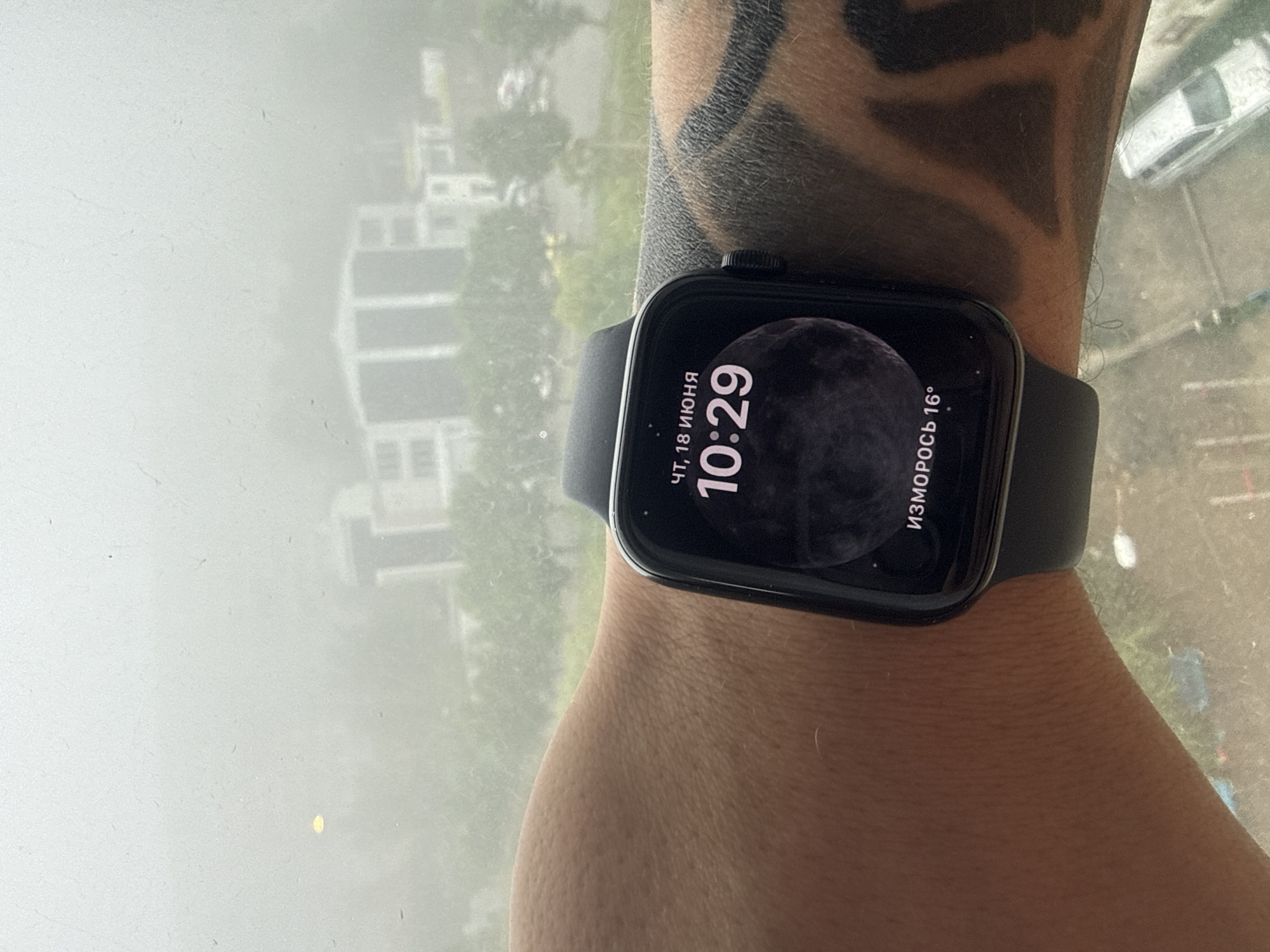 The image shows a close-up of a person's wrist wearing a black smartwatch. The watch face displays the time as 10:29 and the date as Thursday, June 18th. It also shows a weather icon indicating fog and a temperature of 16 degrees Celsius. The background is out of focus, but appears to be an urban environment with buildings and trees visible through a window, possibly on a misty or foggy day. The location is identified as Vladivostok, Russia, although this information is not visually present in the image itself and must be inferred from external context. The person's arm has a tattoo visible on the right side.
