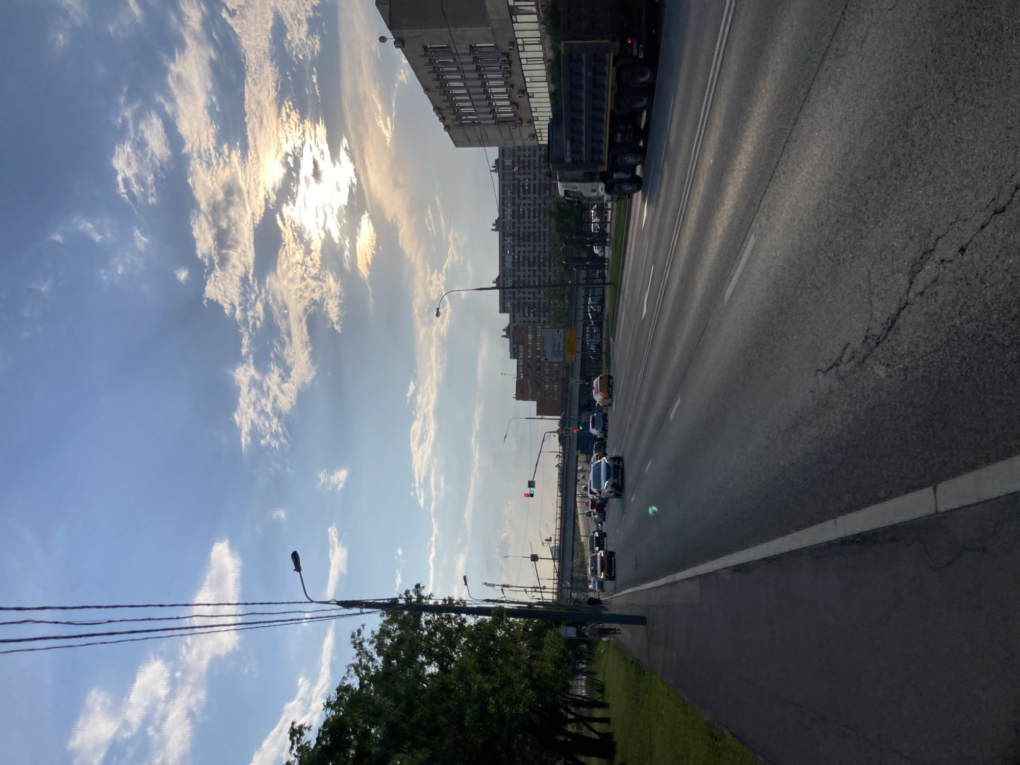 The image depicts a wide, multi-lane asphalt road in Moscow, Russia, with a sidewalk and a grassy area on the left. Overhead power lines stretch across the sky, which is partly cloudy with the sun partially obscured by clouds, creating a dramatic, late afternoon or early evening light. Several vehicles, including cars and a large dump truck, are traveling on the road. In the background, there are multi-story buildings characteristic of urban architecture in Moscow, including a large, gray, modern building with many windows. An overpass or bridge spans the road in the distance, with traffic lights visible. A few pedestrians are walking on the sidewalk. The overall scene conveys a typical urban commute during a transitionary period of the day, with elements of both natural and man-made environments present.