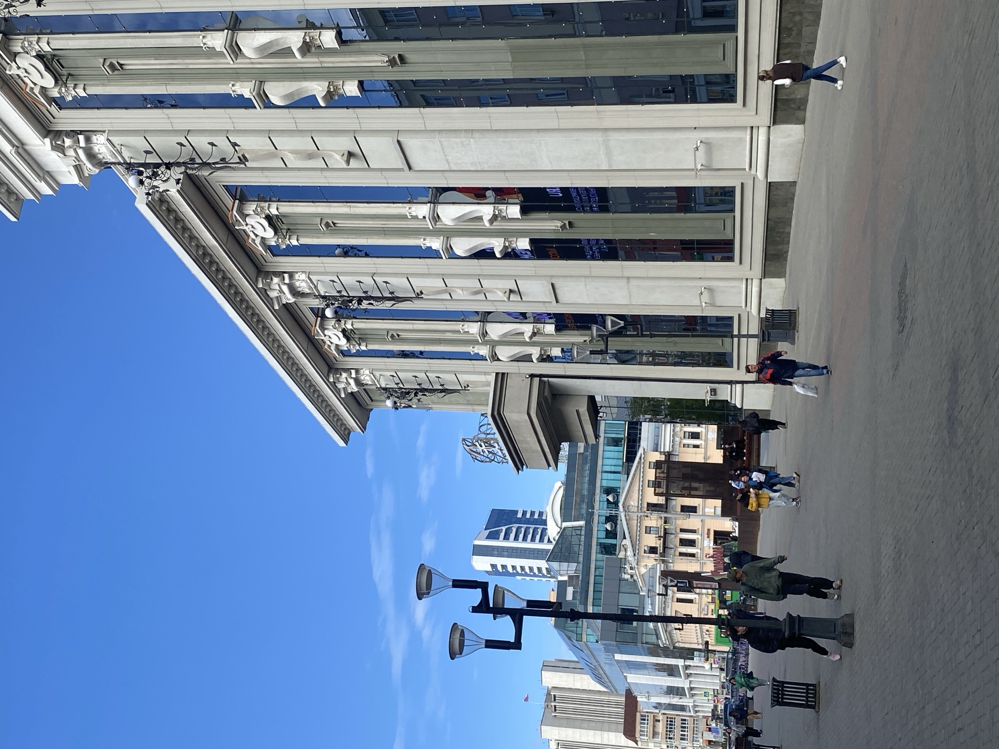 The image shows a street scene in Yekaterinburg, Russia. The sky is clear blue. A large, ornate building with columns and decorative elements occupies the left side of the image. Its windows reflect the sky. On the right, the street pavement stretches into the distance, occupied by several pedestrians. They are walking in different directions. Street lamps are positioned along the street. In the background, several buildings of varying heights can be seen.