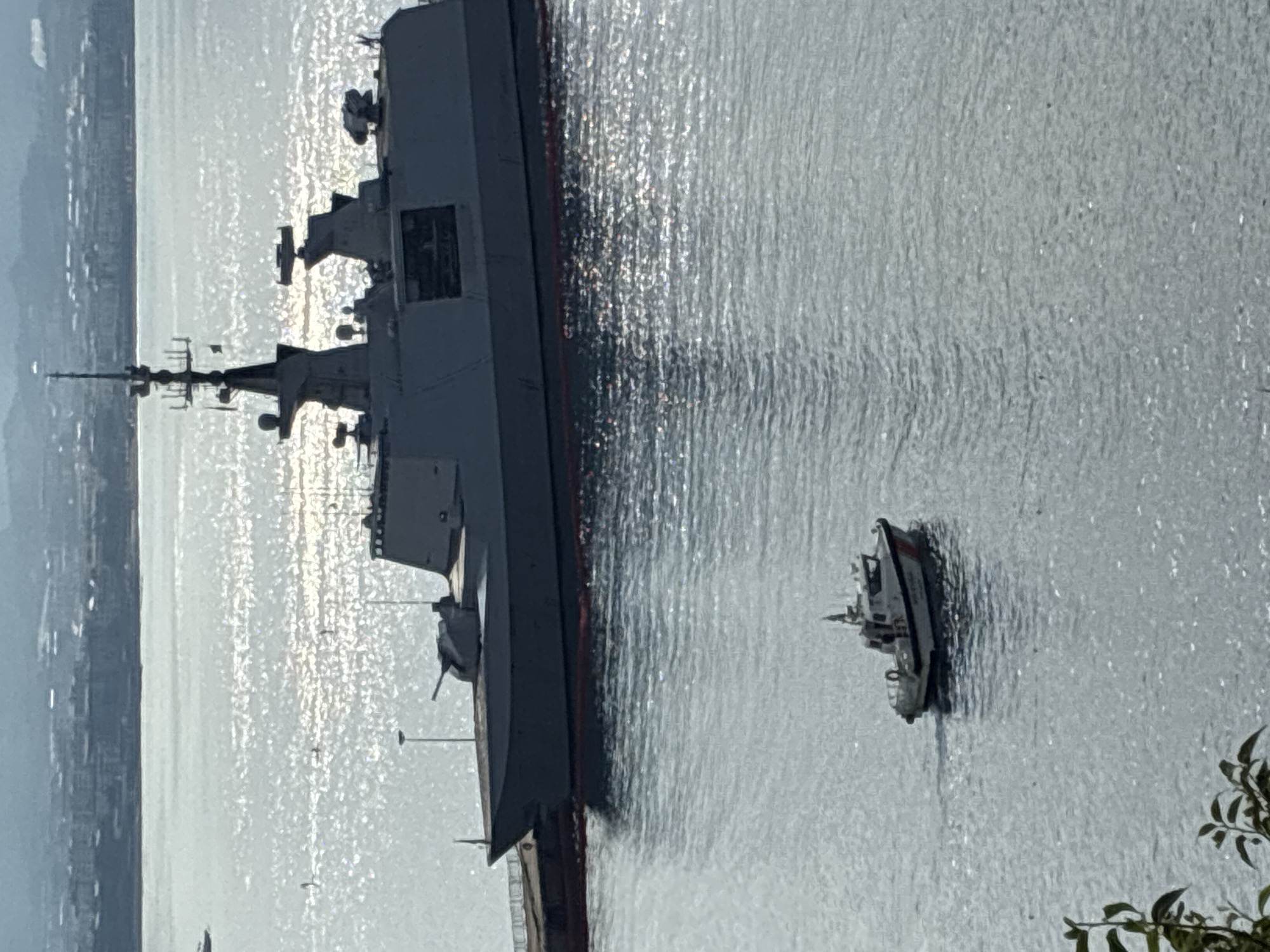 A modern gray naval ship is docked at a pier, its silhouette sharp against the sparkling water. A smaller white and orange patrol boat is navigating the water in front of the larger vessel. In the background, a city skyline with buildings and hills is visible under a hazy sky. The image appears to be taken from a higher vantage point, possibly from a hillside overlooking the harbor.