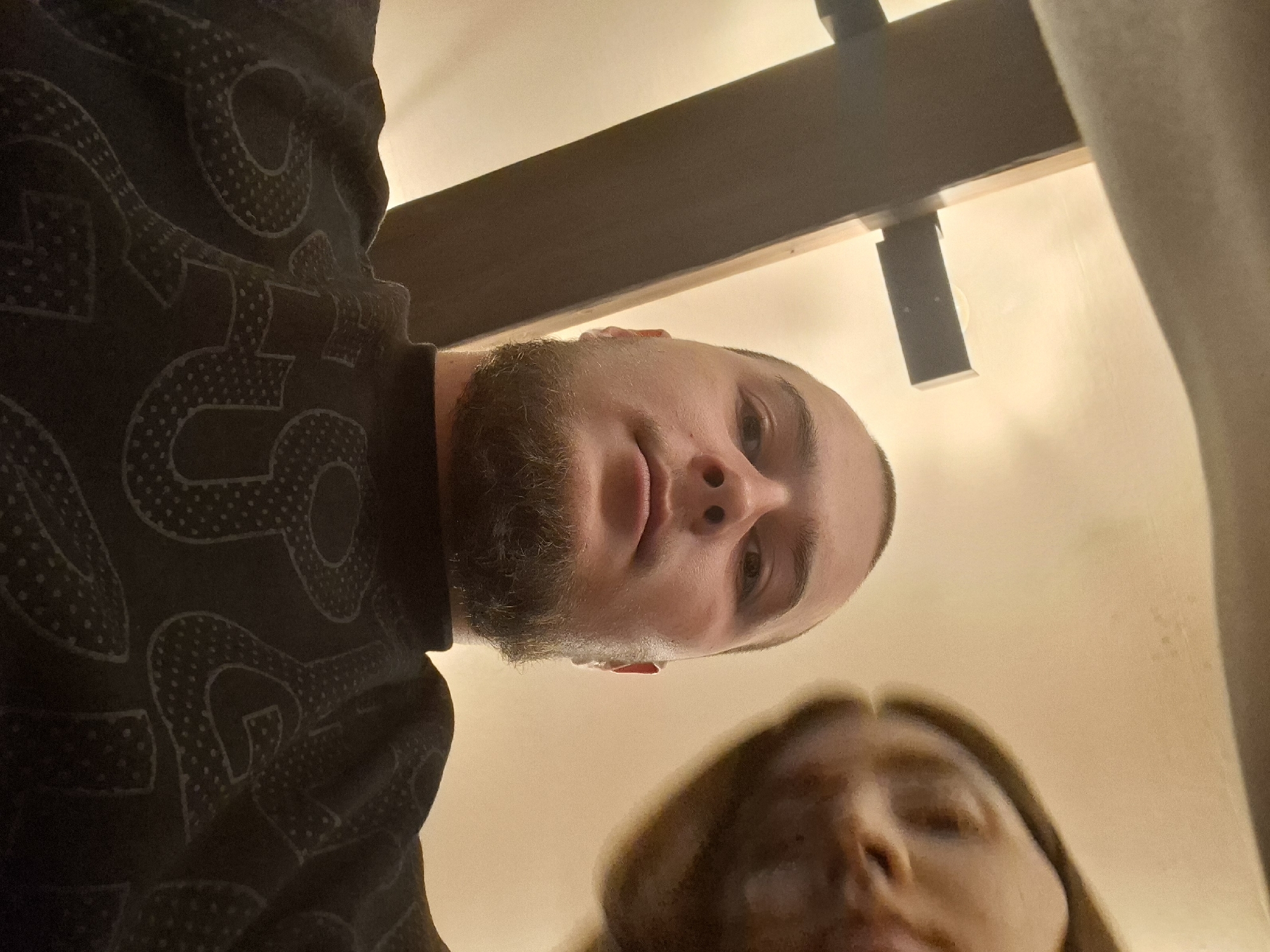 The image shows two people from a low angle, likely taken by one of them. The person in the upper portion of the image is a male with a beard, fair skin, and short hair. He is wearing a dark-colored shirt with a light-colored pattern. The person in the lower part of the image appears to be a female with light-colored hair. Behind the male, a wooden structure is visible. The background includes what appears to be a wall or curtain. The exact location in Pleven, Bulgaria, is not discernible from the image.