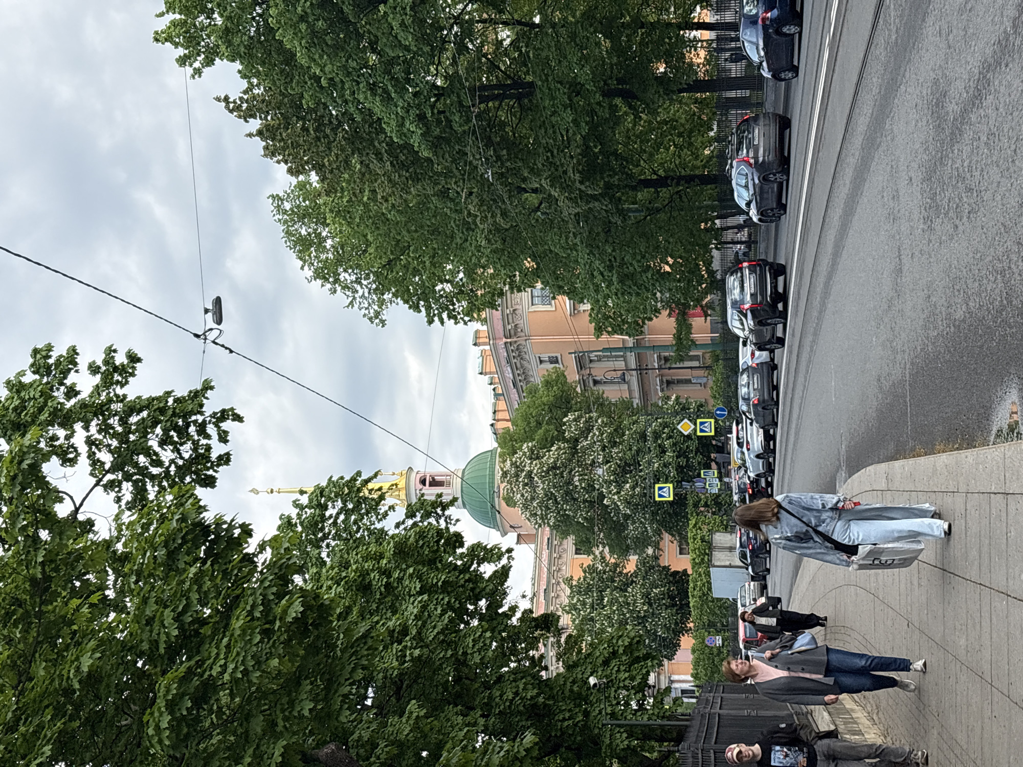 Here is a forensic description of the image:
The image depicts a street scene in Saint Petersburg, Russia. The sidewalk on the right side of the frame is occupied by pedestrians. A woman in a light blue outfit carries a large white shopping bag. Further down the sidewalk, another woman in a gray jacket and blue jeans is also walking. A man with a cap, wearing a t-shirt, and gray pants is at the front.
A row of parked cars lines the street. Street signs with pedestrian crossing symbols are visible near the cars. The road includes tram tracks.
Trees line both sides of the street. Through the trees on the left, the dome and spire of a building are visible. The building's dome is green and its spire is gold. Another building behind the trees shows orange and red tones. Power lines and a street light run over the road, partially obscured by the trees.
The sky overhead appears overcast.