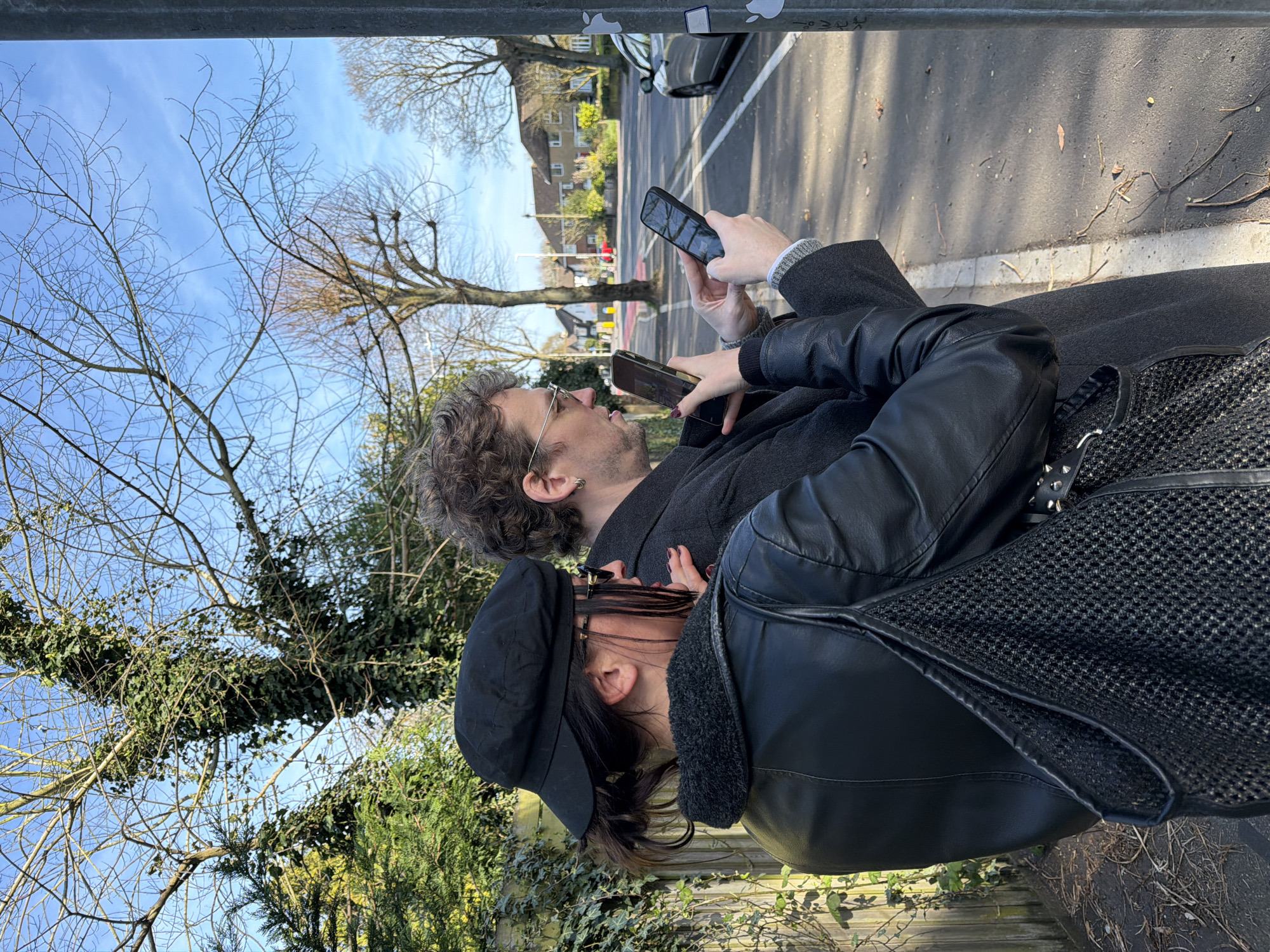 Two individuals, a man and a woman, are standing outdoors near a paved street in Folkestone and Hythe District, United Kingdom. The man, wearing glasses and a dark coat, is holding a smartphone at eye level. The woman, positioned slightly behind him, is wearing a black cap and a leather jacket with a mesh bag. She also appears to be holding a smartphone. They are both facing forward, presumably looking at their respective phones. The scene includes trees with bare branches and ivy, residential buildings across the street, a parked car, and street markings. The sky is visible through the branches and is partially clear with some clouds. A gray structure or pole is in the foreground, partially obscuring the top of the image.