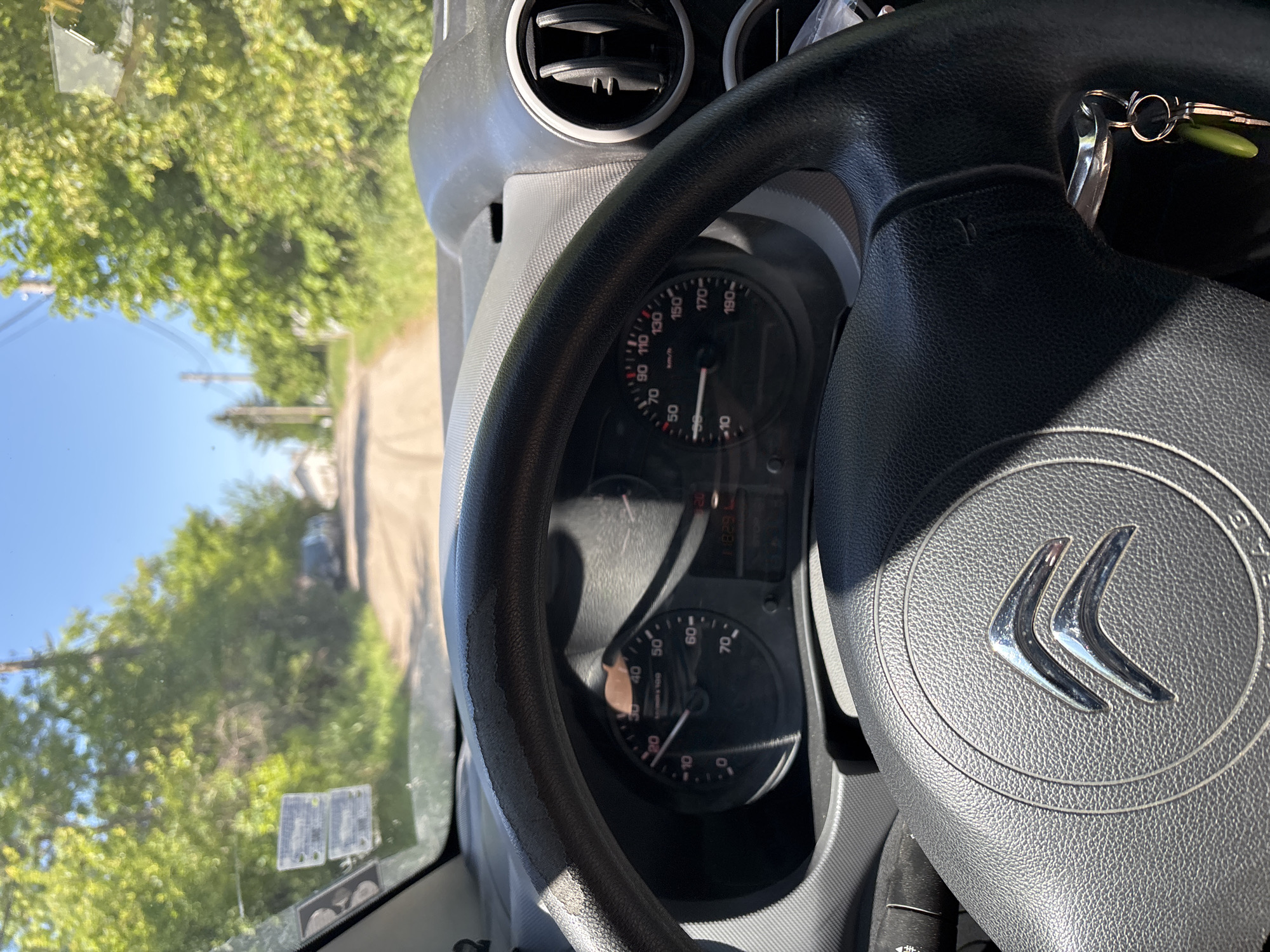 A view from inside a car, likely a Citroën, featuring the steering wheel with the Citroën logo prominently displayed. The dashboard is visible, showing the speedometer, which indicates a speed just above 0 km/h. Through the windshield, a road is seen lined with trees, possibly located in a rural or suburban area. Given the location listed in the prompt, this scene may depict a street in Pojarevo, Bulgaria. The car appears to be stationary.