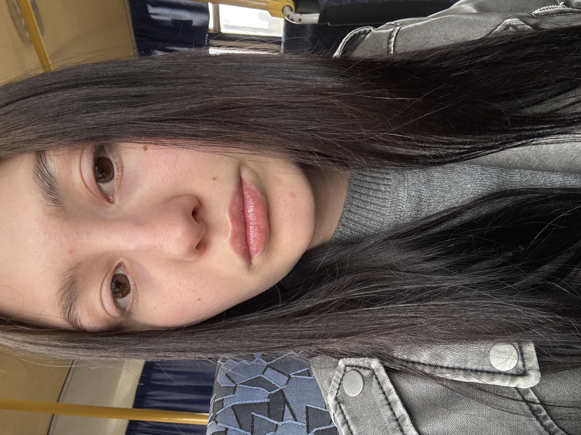 A selfie of a young woman with dark hair and light skin. She is wearing a grey sweater and a silver jacket with large, round buttons. The background suggests she is on public transportation, likely a bus or train, with a yellow rail and blue seats visible. The location is uncertain, but the information indicates it may be in the city of Фоки, Russia.