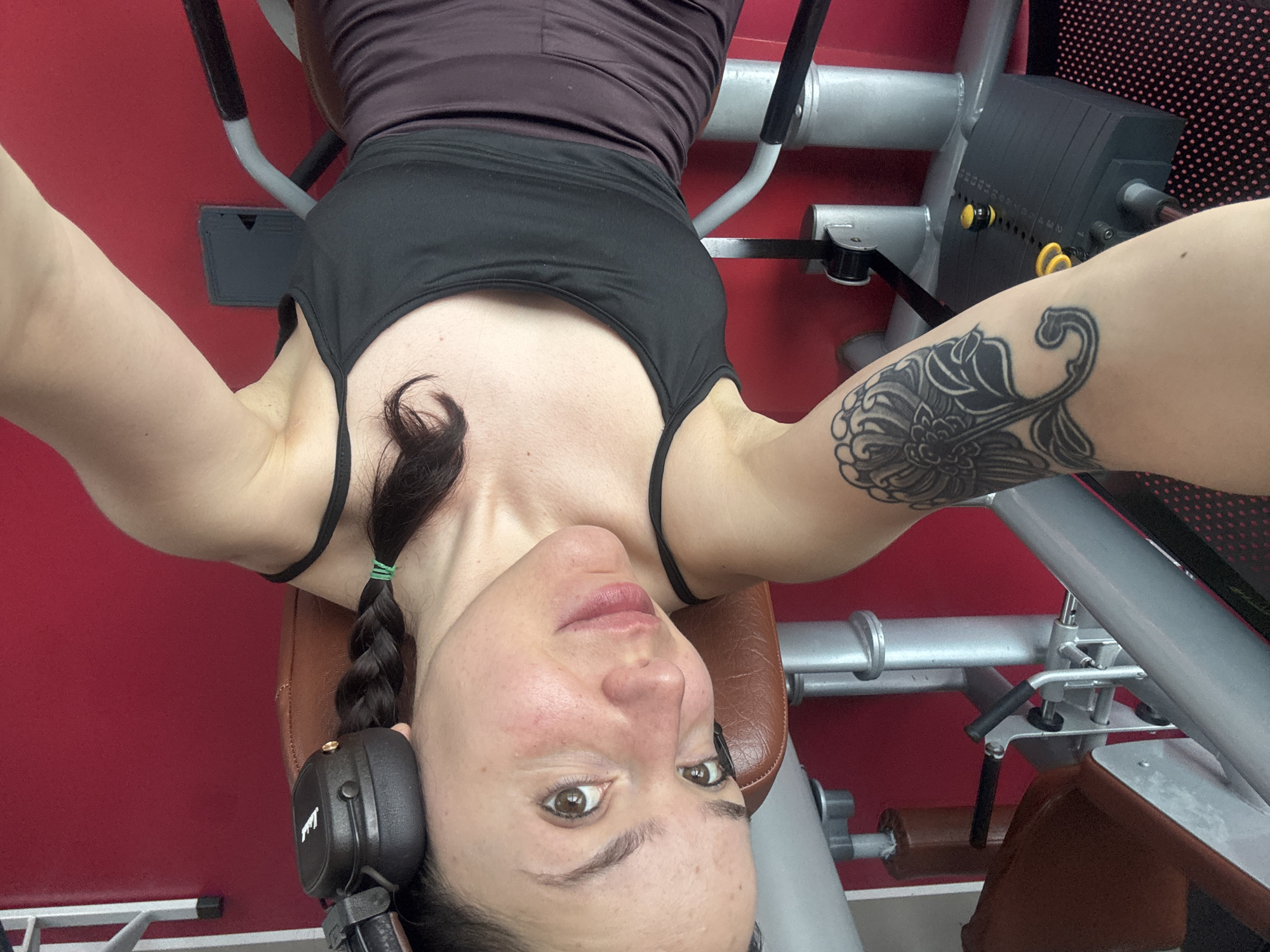A woman with a braided hairstyle and a black tattoo on her right arm is lying on a brown bench in a gym. She is wearing a black tank top and a purple tank top underneath. Headphones are partially visible on her left ear. Exercise equipment surrounds her, including a weight stack and various metal bars and supports. The background shows a red wall. The location is potentially in Burgas, Bulgaria.