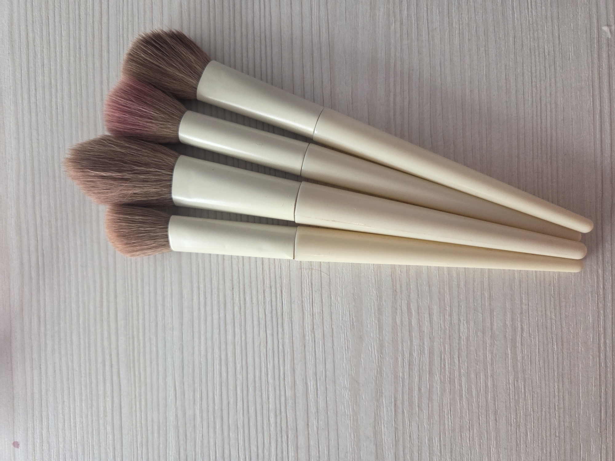 Four makeup brushes with light beige handles and brown bristles are arranged diagonally on a light-colored surface with a wood grain texture. One brush has some pink pigment on the bristles. No people are visible. The location Yug, Russia is irrelevant to the image's content.