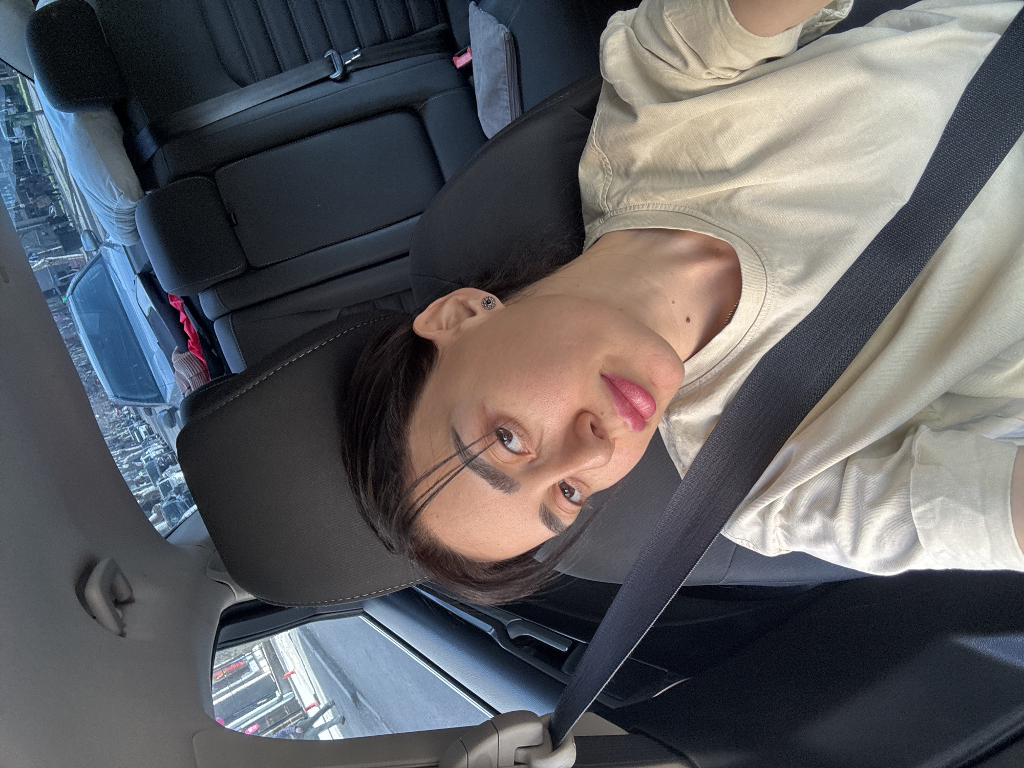 The image is a close-up selfie of a person wearing a seatbelt while sitting in a car. The person has dark hair and light skin, and is wearing a light-colored shirt. The car's interior is black leather. Through the window, a cityscape can be seen, which appears to be in Tbilisi, Georgia, judging from the architecture and landscape.